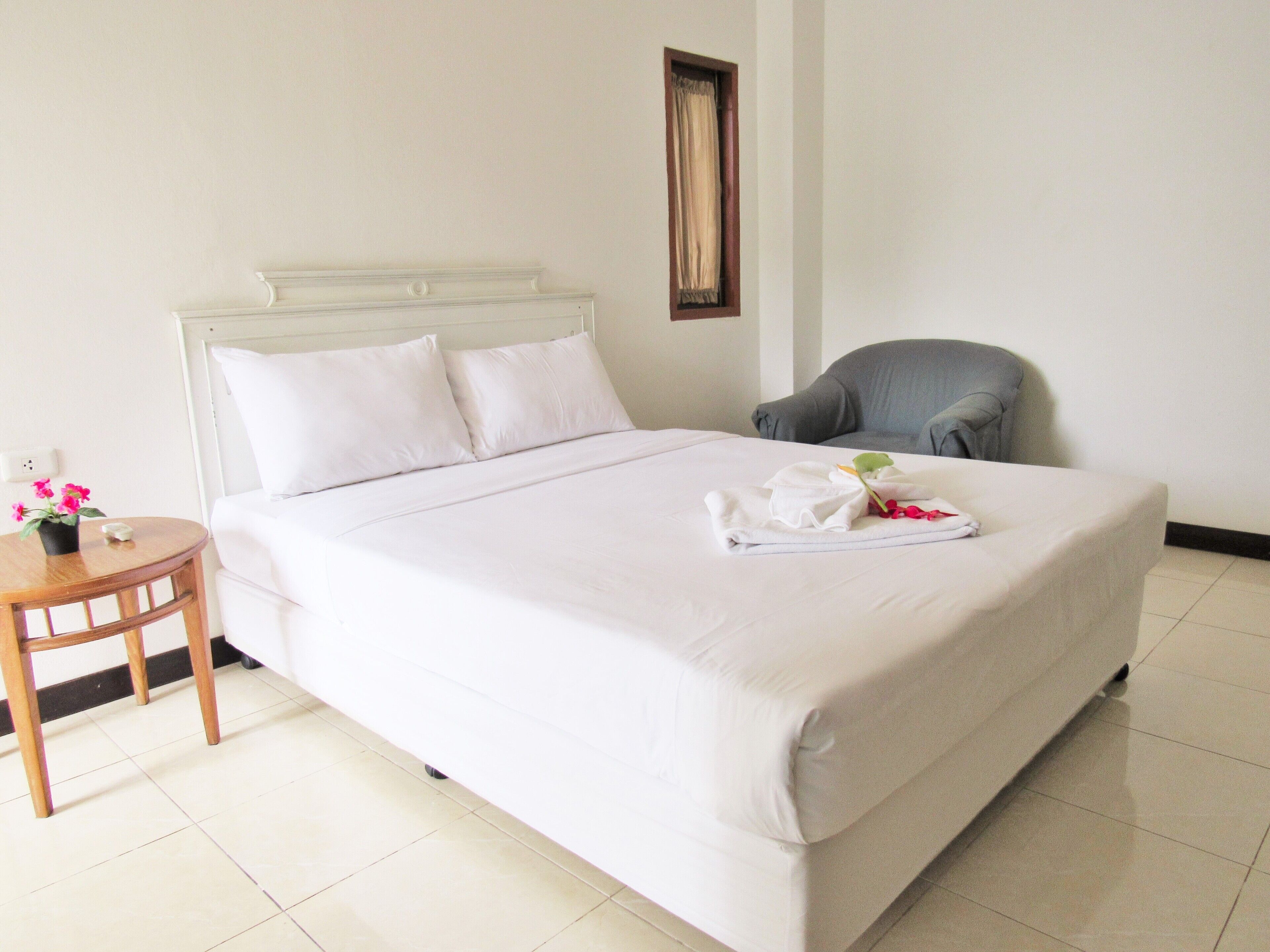 A neatly made bed with white linens, a small round table with a potted plant, and a grey armchair in a simple, clean room.