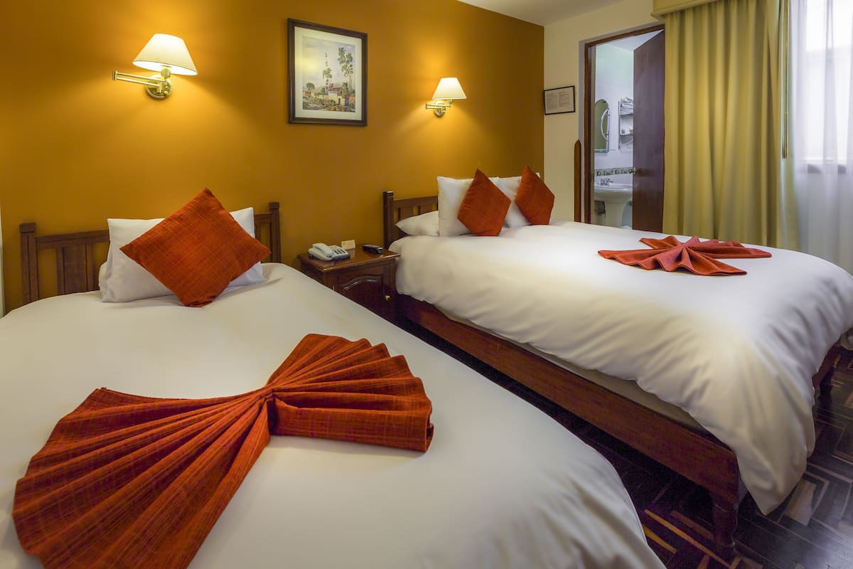 A hotel room with two beds, each with white linens and orange pillows. There are wall-mounted lamps and a painting on the wall.