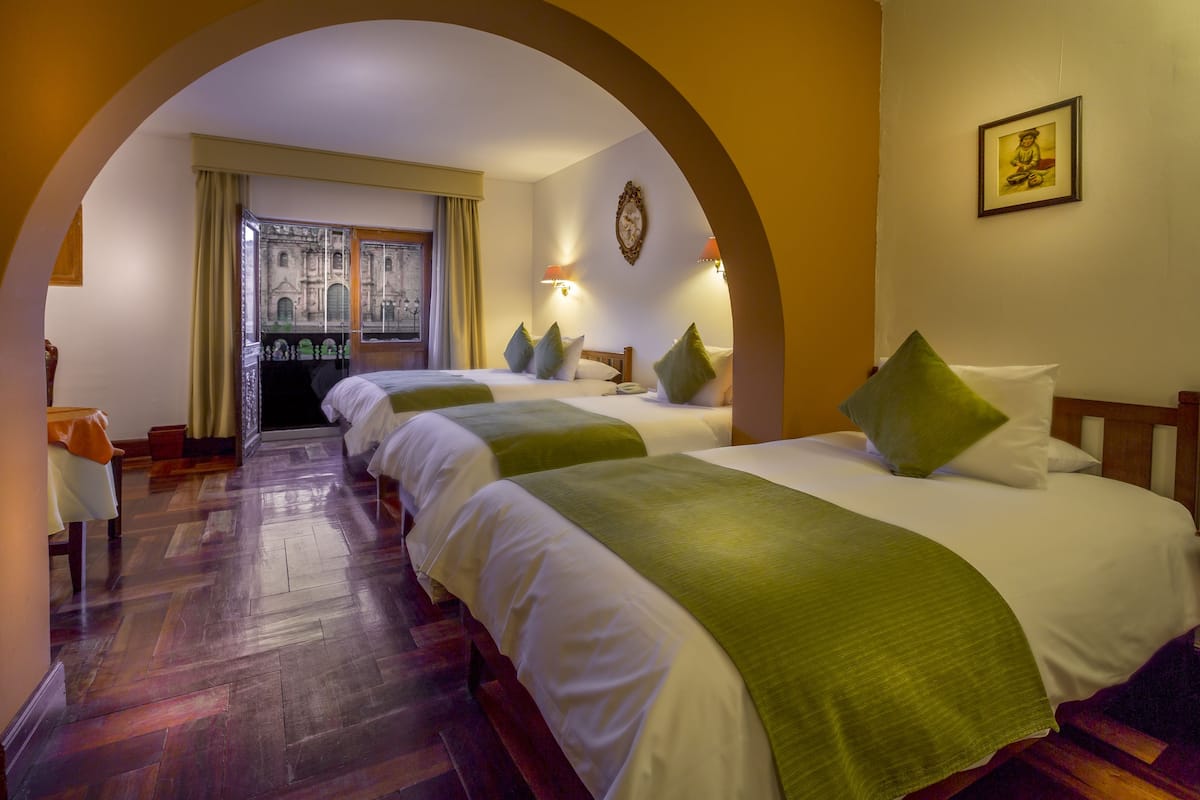A hotel room with three beds, wooden flooring, and a view through an archway.