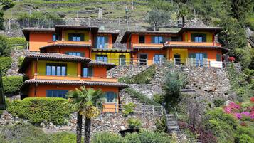 Un edificio de varios pisos con fachada de piedra y tejas de terracota, con balcones y una escalera que sube por la colina.