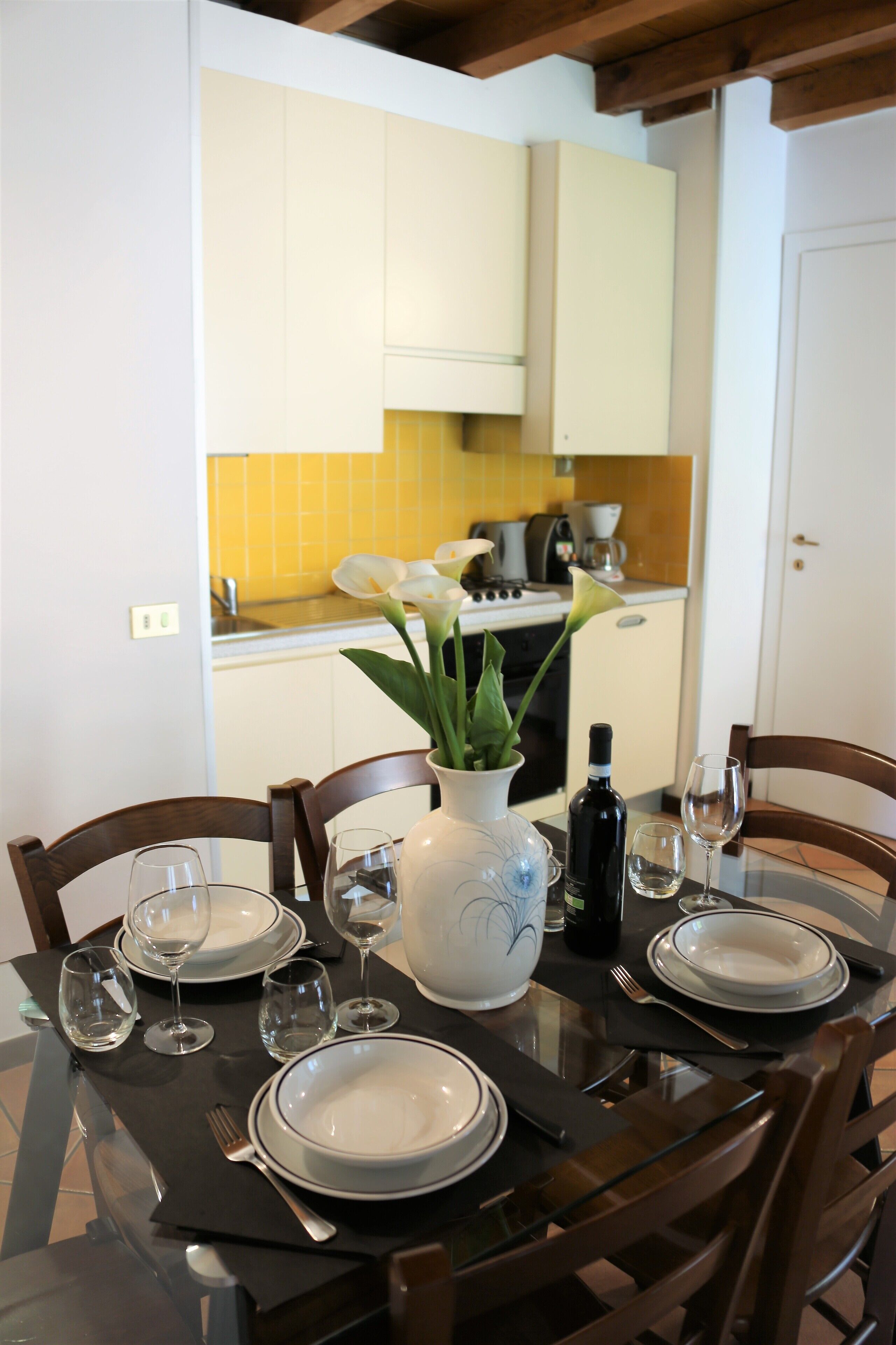 A dining table set with plates, glasses, cutlery, a vase with flowers, and a bottle of wine in a modern kitchen with white cabinets and a yellow backsplash.