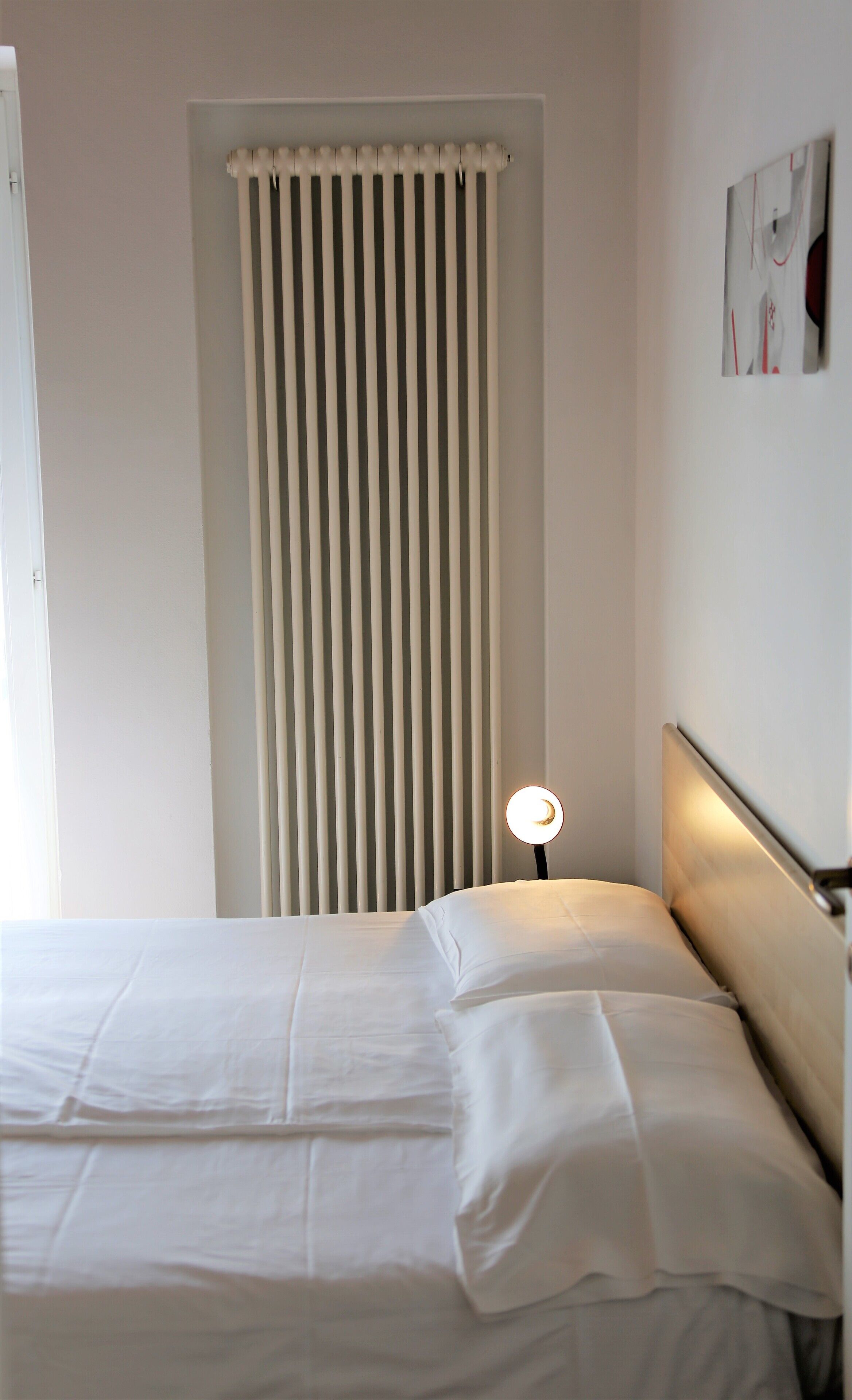 Un dormitorio con una cama, una mesita de noche con lámpara, una ventana con persianas verticales y una pared con un cuadro enmarcado.