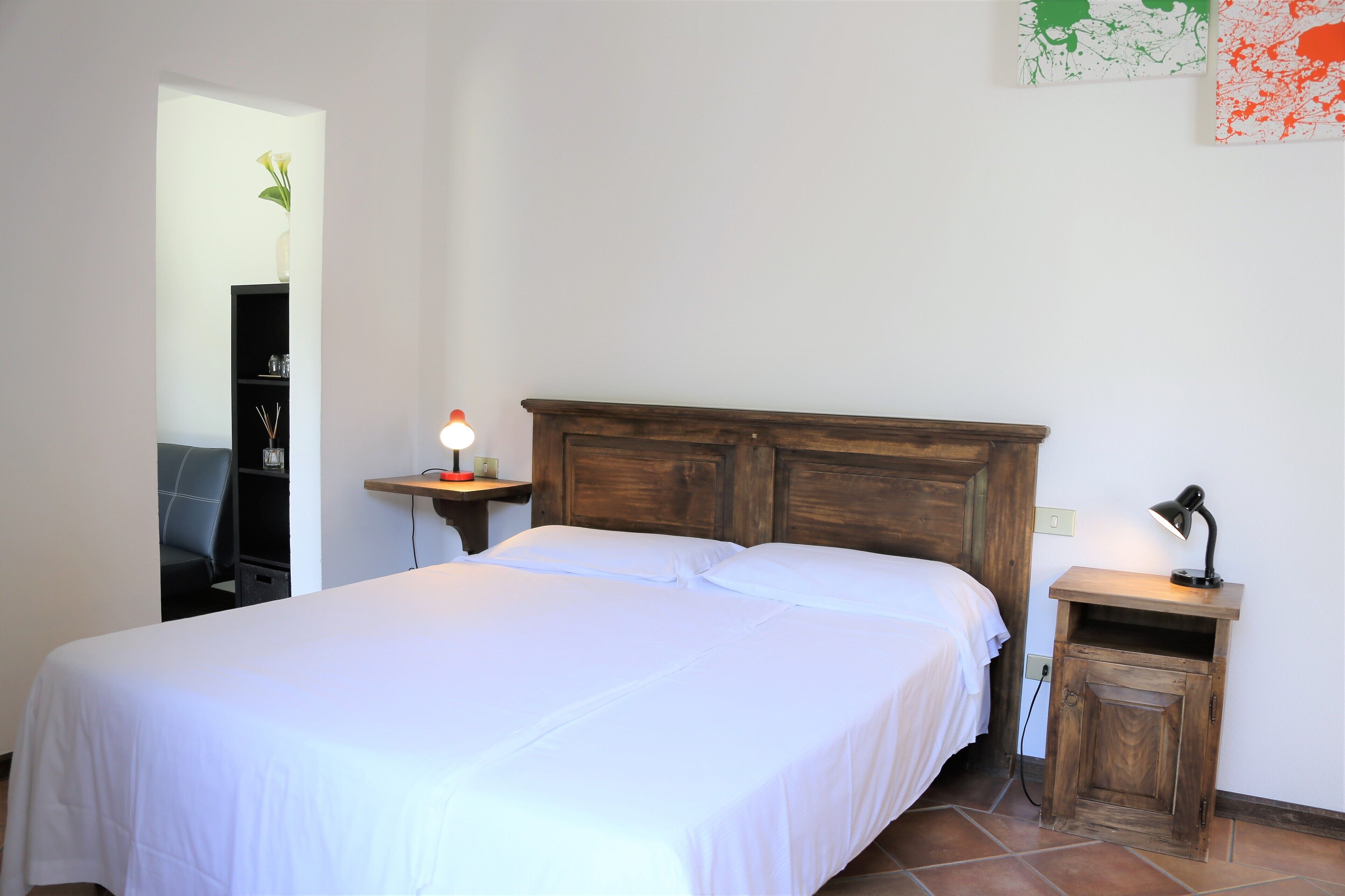A neatly made bed in a room with a wooden headboard, a small bedside table with a lamp, and a wall-mounted shelf with a vase.