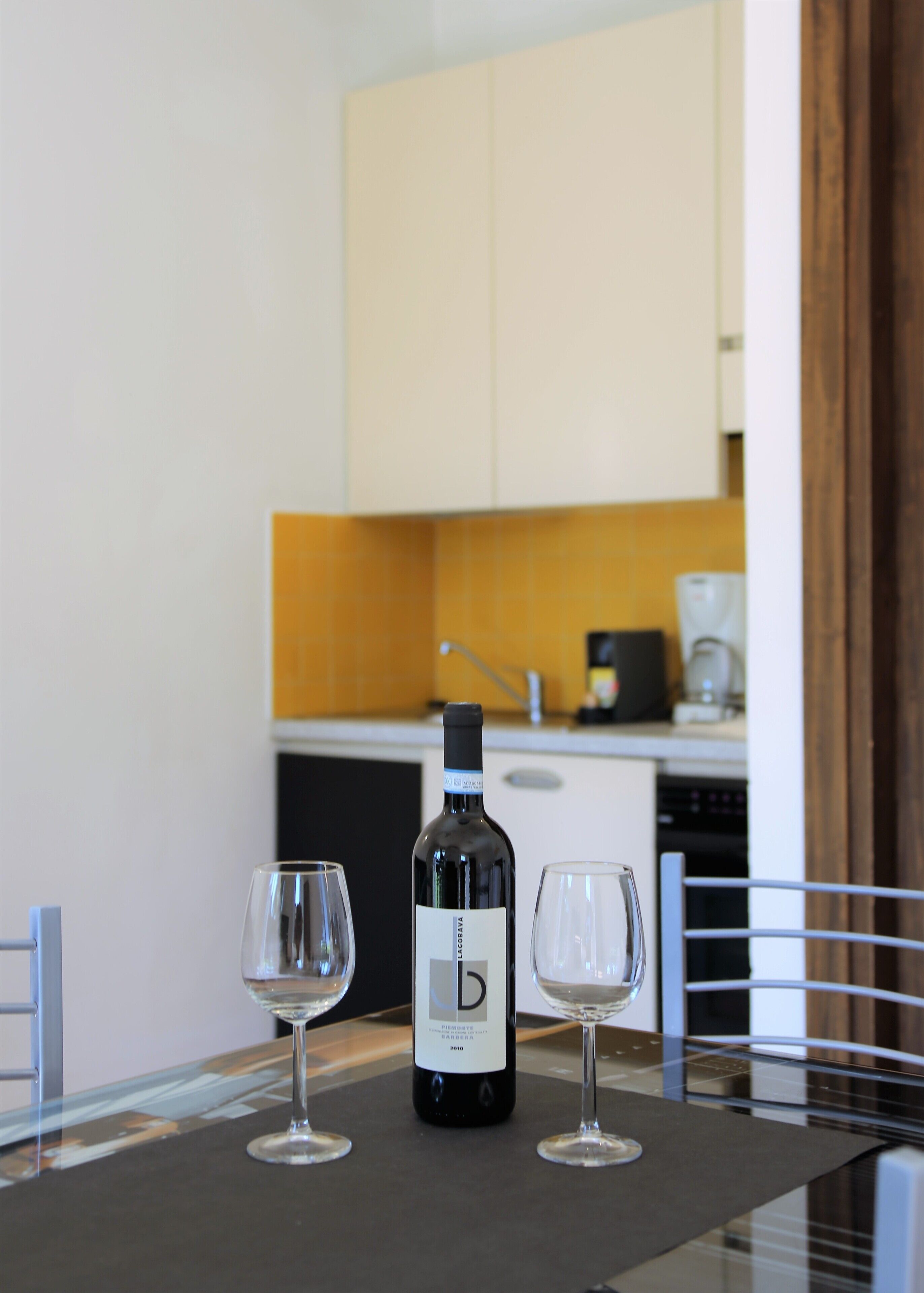 Una mesa de comedor con una botella de vino y dos copas, una cocina al fondo y un marco de puerta de madera.