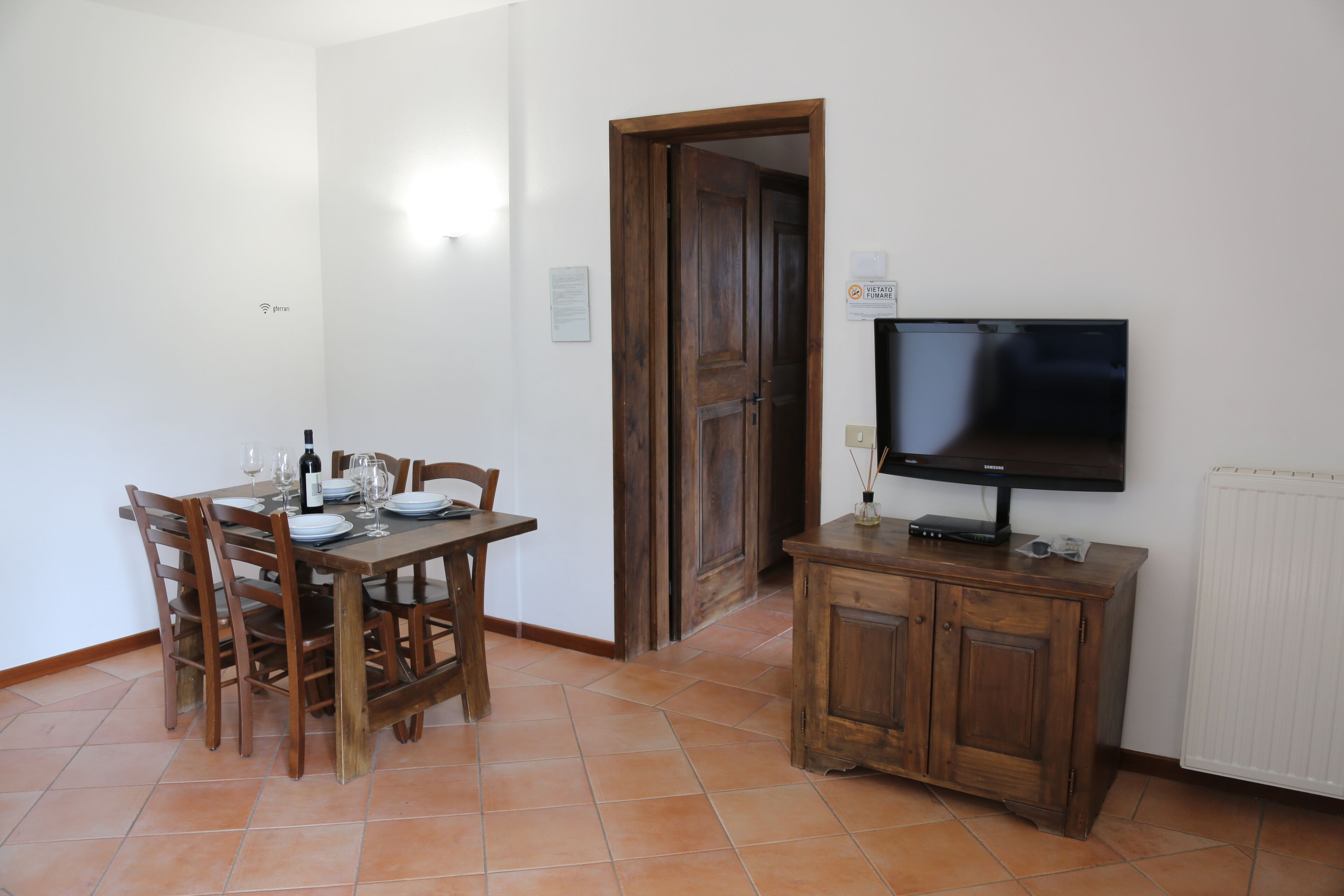 Uma área de refeições com uma mesa de madeira posta para quatro pessoas, uma televisão sobre um móvel de madeira e um radiador branco.