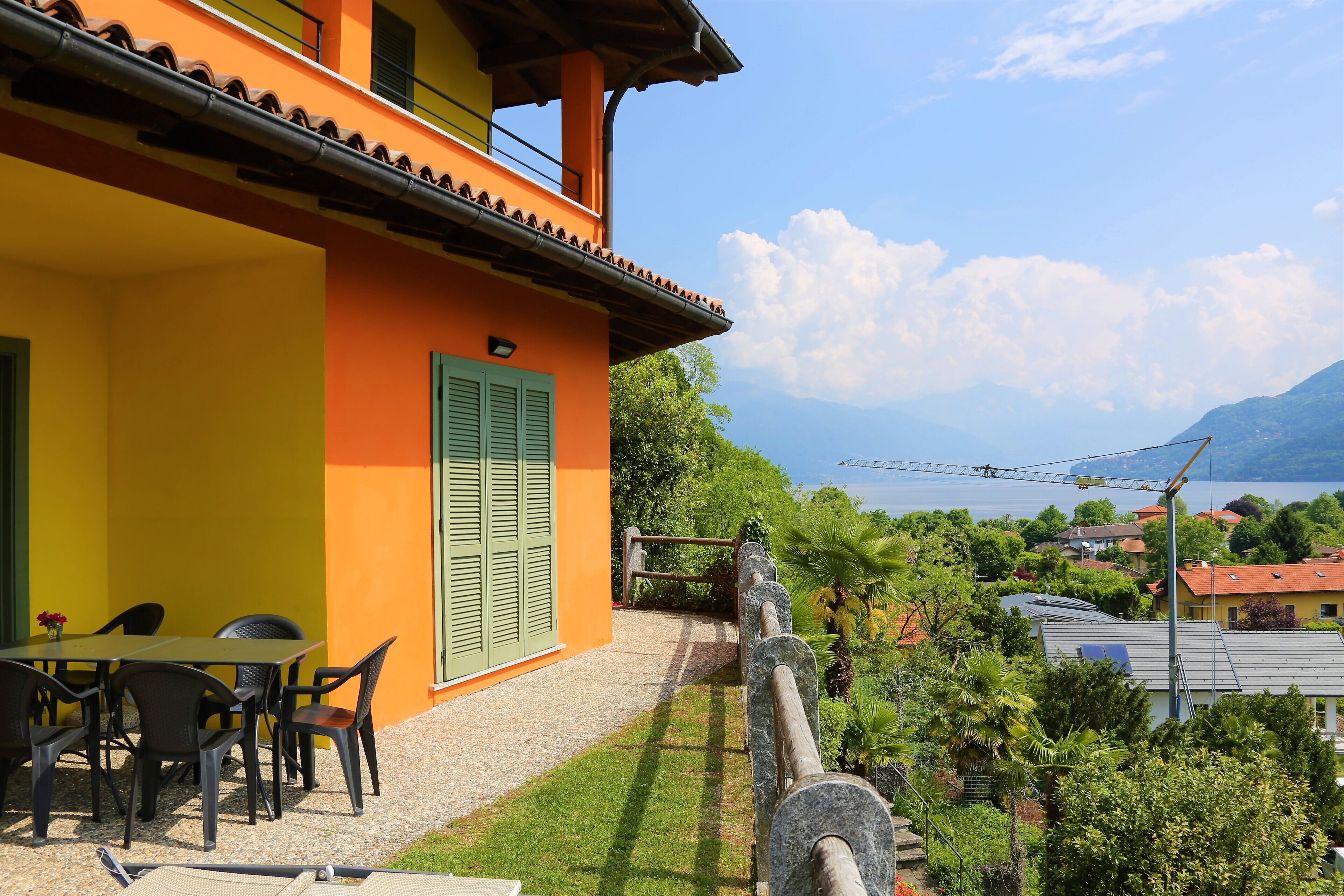 A building with a yellow exterior and green shutters, a patio with outdoor furniture, and a view of a lake and mountains in the background.