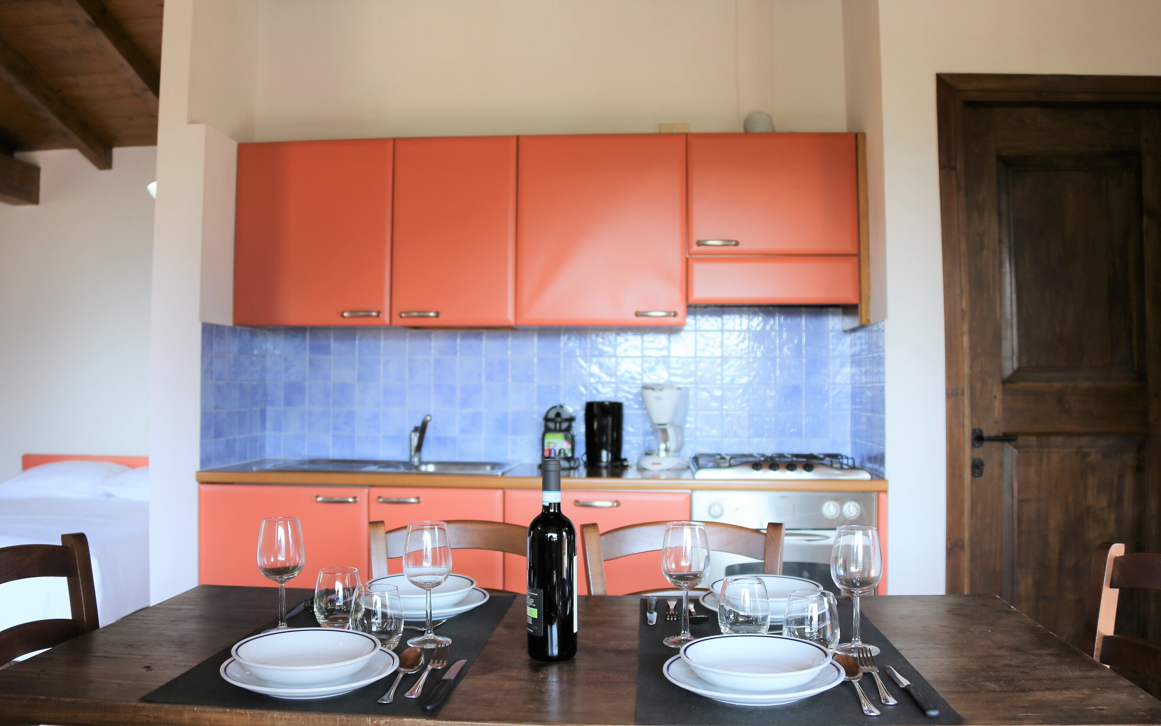 A dining table set with plates, wine glasses, and cutlery in a kitchen with orange cabinets and a blue-tiled backsplash.