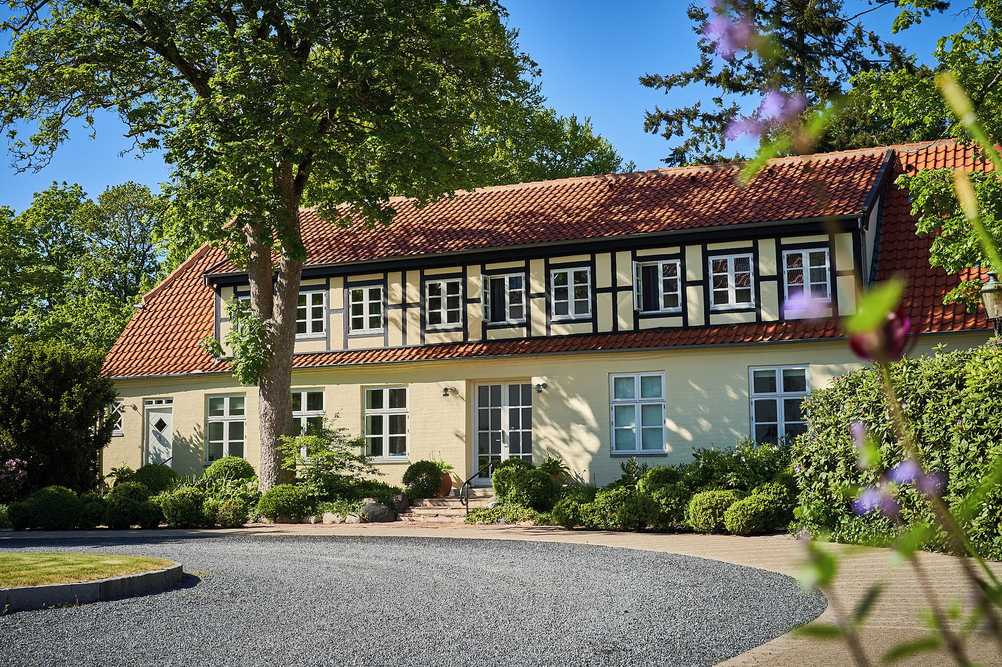 A traditional half-timbered house with a red-tiled roof, surrounded by well-maintained greenery and a gravel driveway.