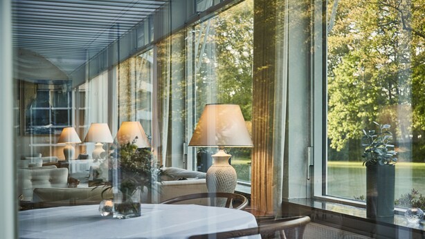 A dining area with a table set for two, a lamp, and a view of greenery outside. - Gl. Skovridergaard (Silkeborg)
