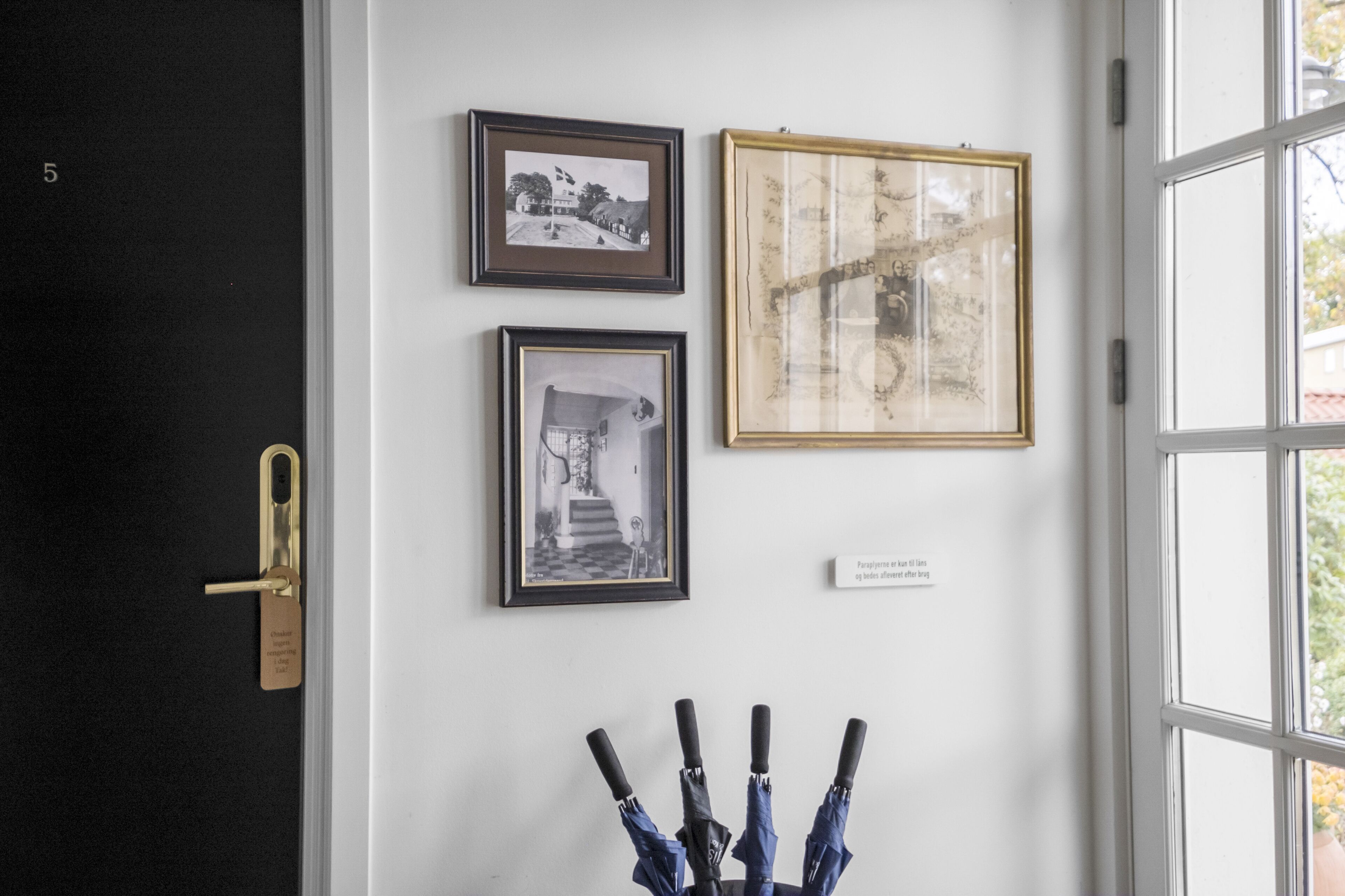 A wall with three framed pictures, a door with a golden handle, and a window with a view of greenery.