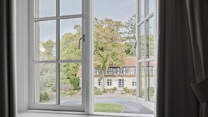 A view from a window with white frames, looking out onto a well-maintained lawn, a building with a traditional facade, and a clear sky. - Gl. Skovridergaard (Silkeborg)