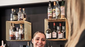 A bar area with shelves of liquor bottles, a bartender pouring a drink, and a customer holding a glass. - Gl. Skovridergaard (Silkeborg)