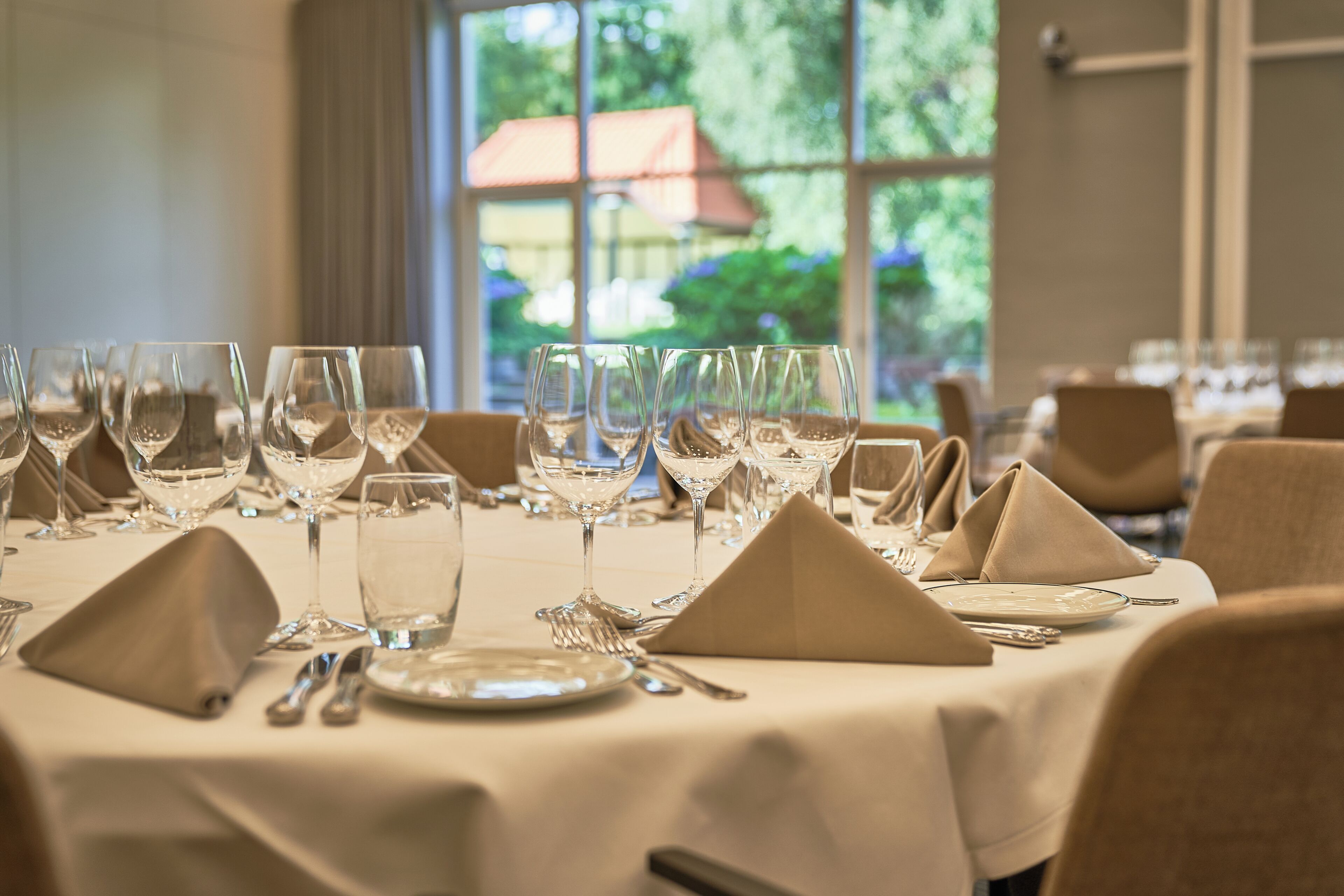 A dining table set with multiple wine glasses, plates, and cutlery, with a view of greenery outside through large windows.