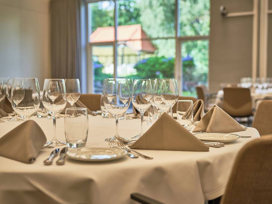 A dining table set with multiple wine glasses, plates, and cutlery, with a view of greenery outside through large windows.