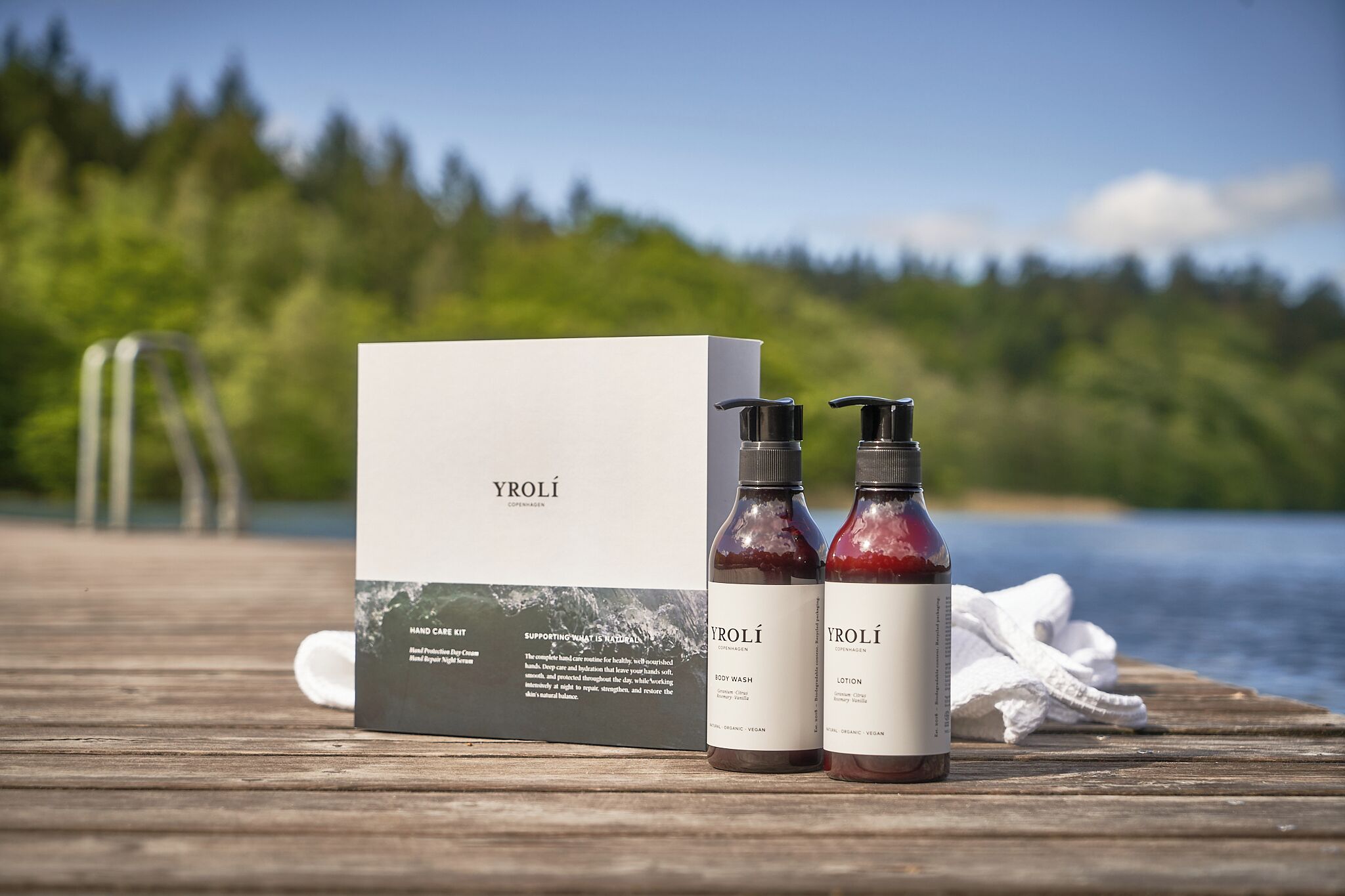A wooden deck with a box and two bottles labeled 'YROLÍ' on a lakeside setting.
