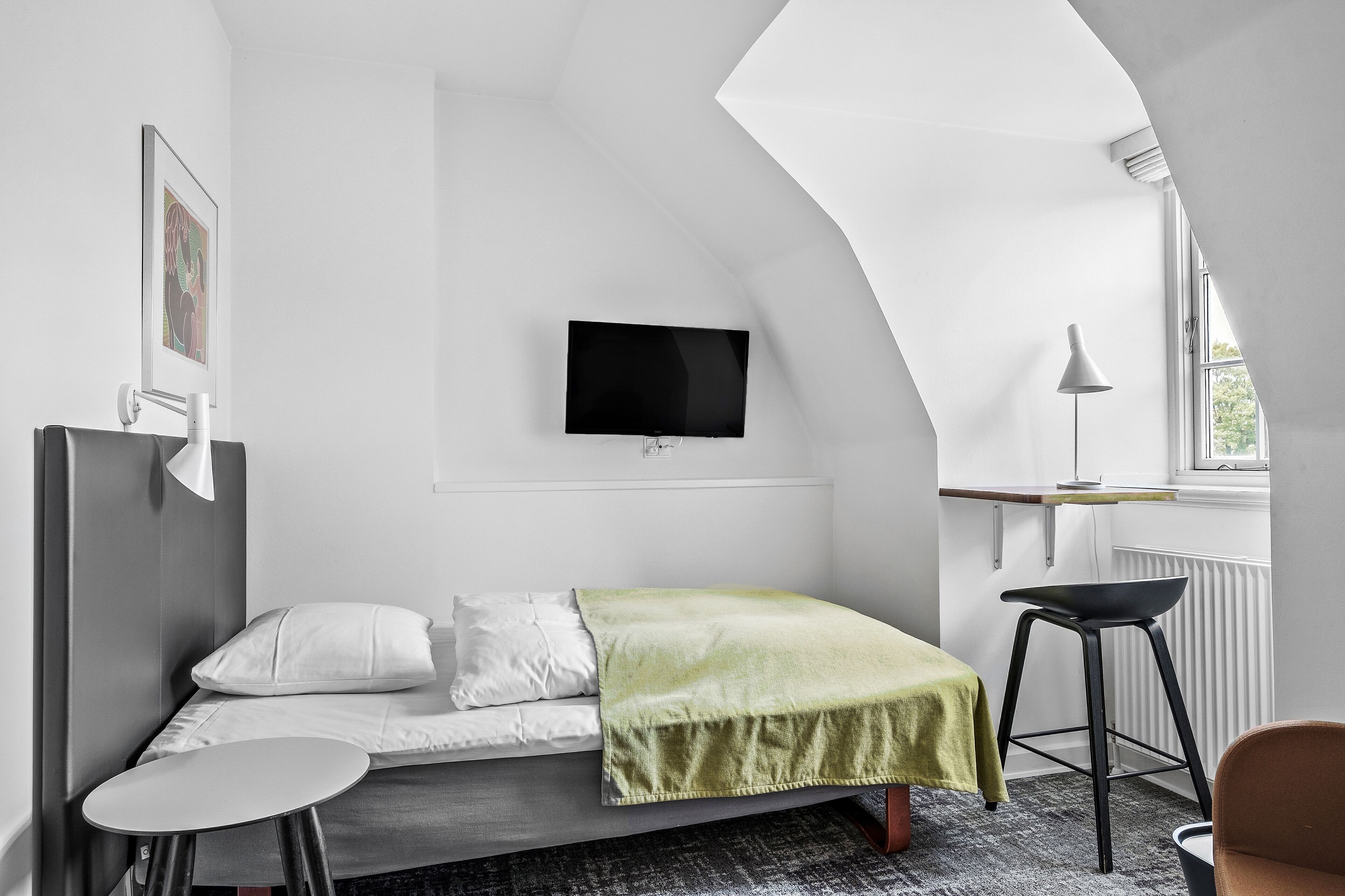 A modern, minimalist bedroom with a bed, a small table, a chair, a TV mounted on the wall, and a window providing natural light.