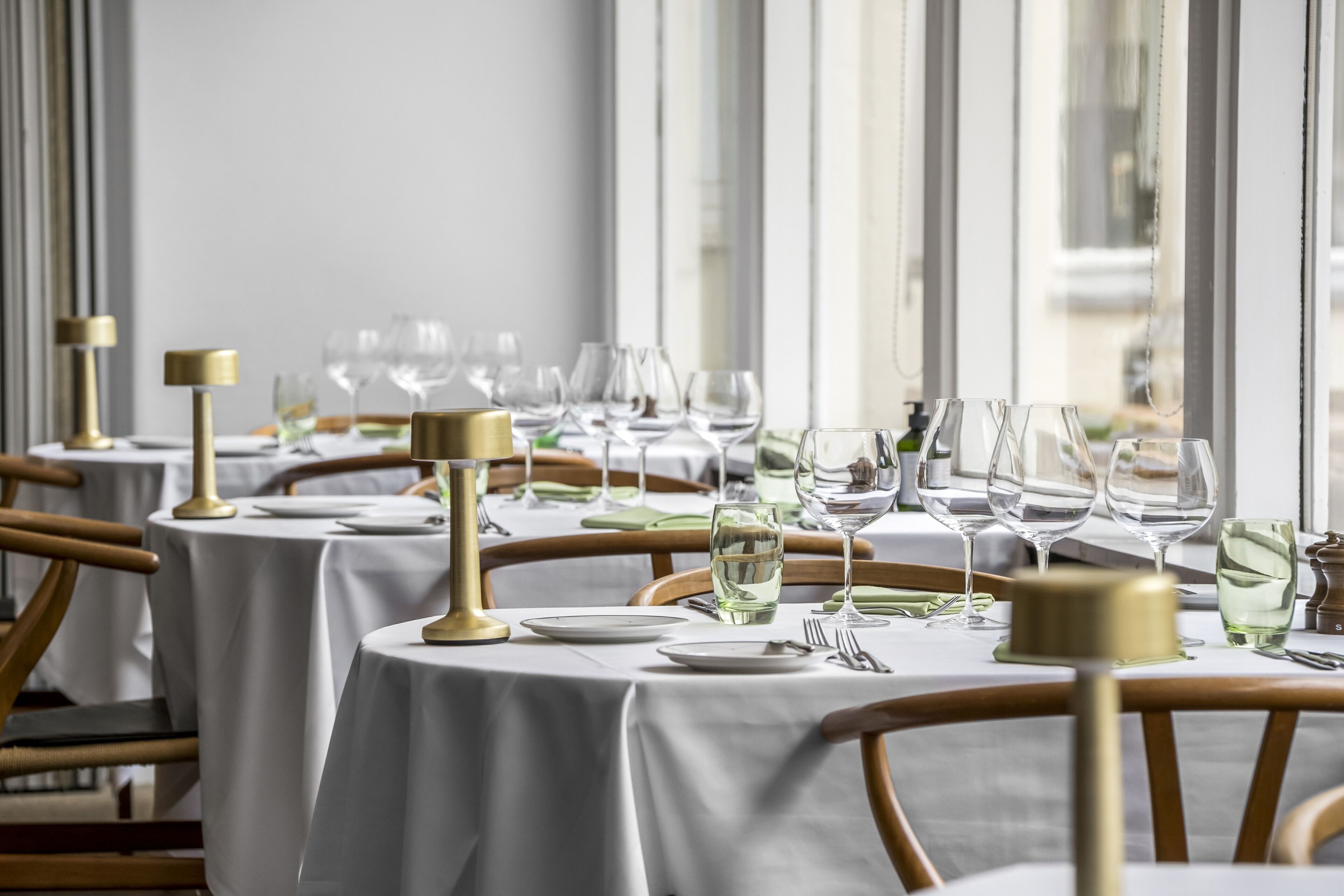 Un restaurant avec de nombreuses tables dressées pour le dîner, chacune étant recouverte d’une nappe blanche, agrémentée de bougeoirs dorés et de verres à vin.