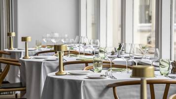 Un restaurant avec de nombreuses tables dressées pour le dîner, chacune étant recouverte d’une nappe blanche, agrémentée de bougeoirs dorés et de verres à vin.