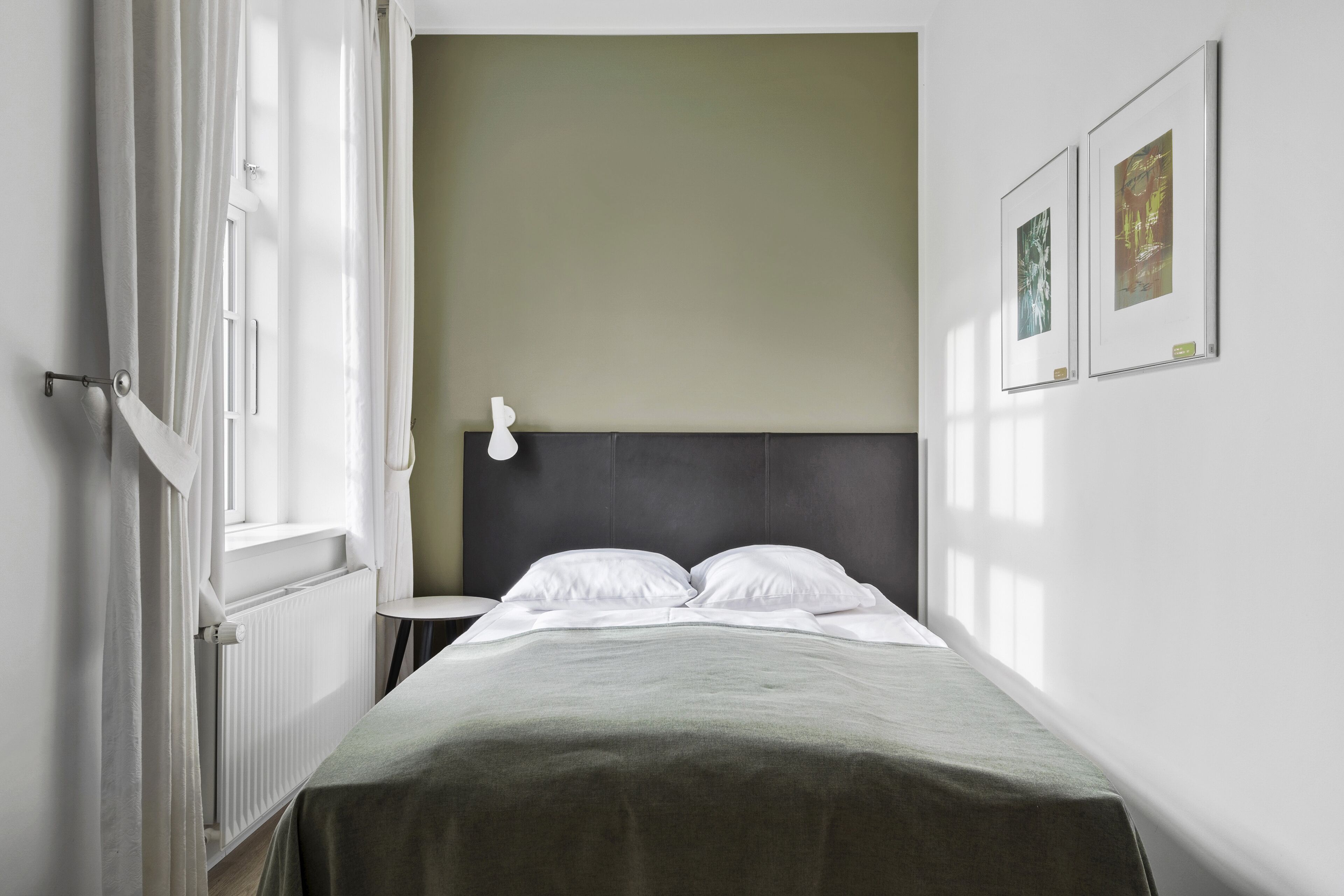 Minimalistinen hotellihuone, jossa on sänky, yöpöytä ja seinään kiinnitetty valaisin.