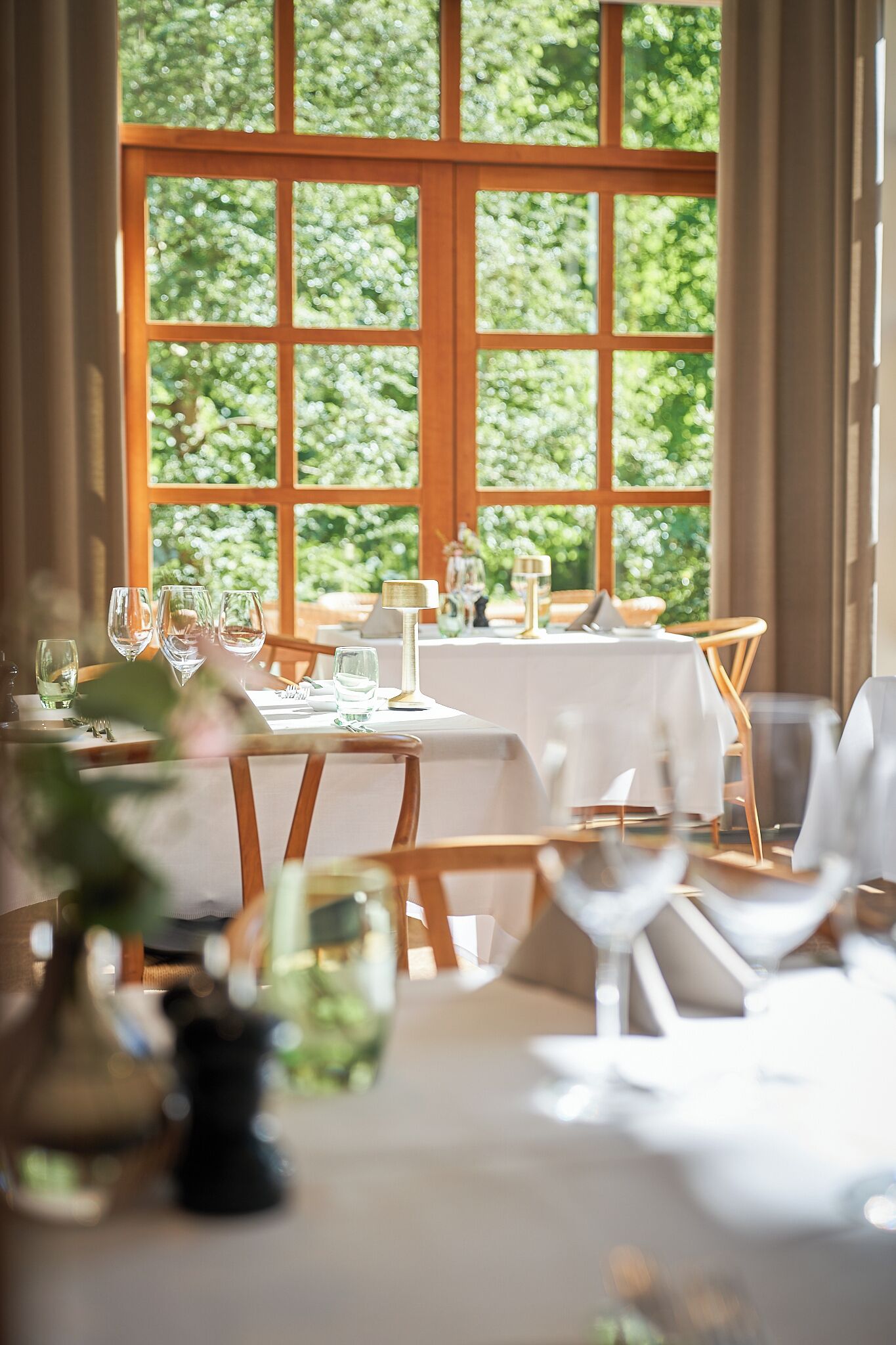 Un comedor con mesas dispuestas para dos, una ventana que ofrece vistas a zonas verdes, y un jarrón con flores sobre la mesa.