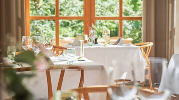 Un comedor con mesas dispuestas para dos, una ventana que ofrece vistas a zonas verdes, y un jarrón con flores sobre la mesa.