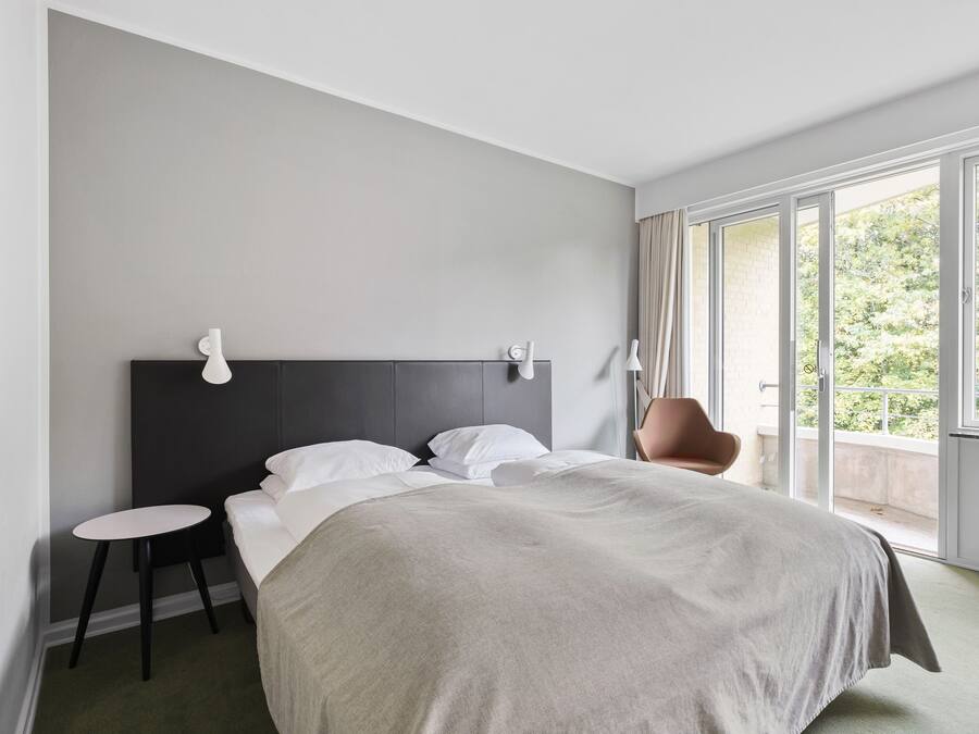 A modern hotel room with a large bed, a small side table, a chair, and a view of greenery through large windows.