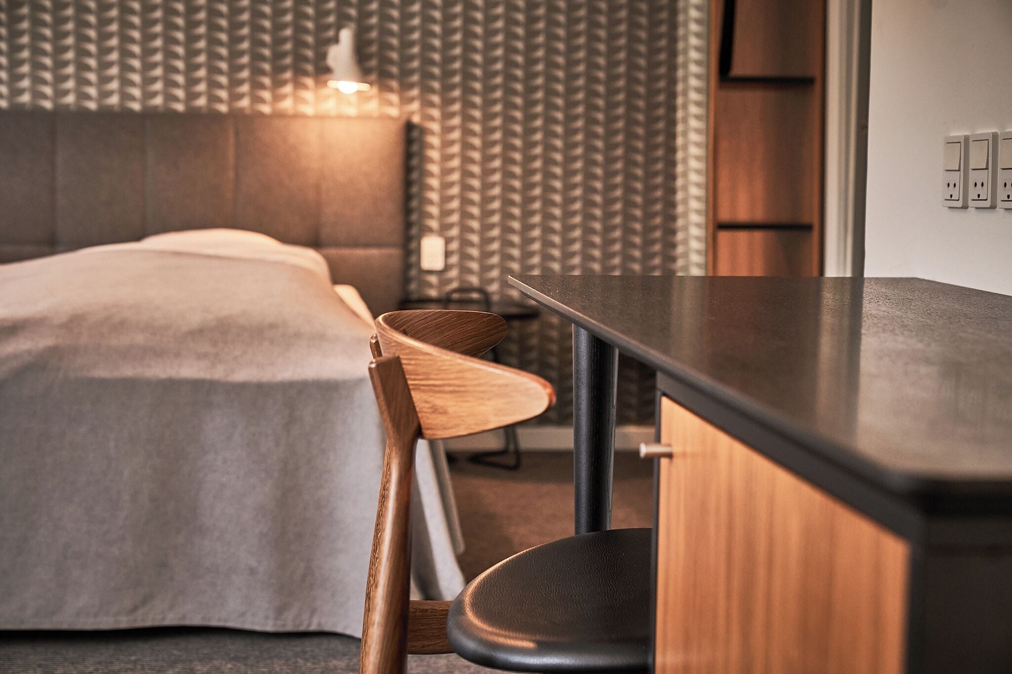 A modern room with a bed, a wooden chair, a desk, and a patterned wallpaper.
