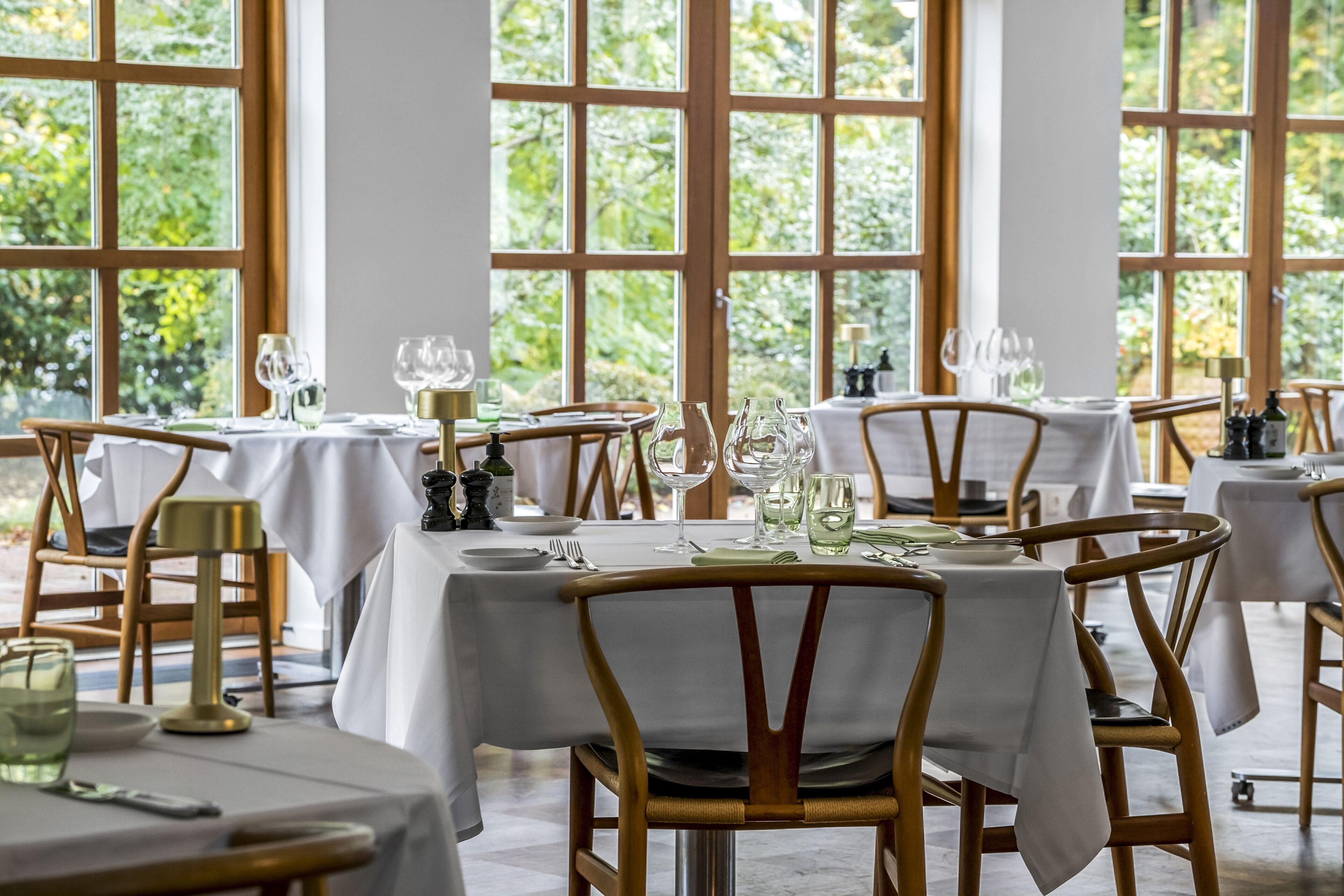 A restaurant with tables set for dining, large windows, and a view of greenery.
