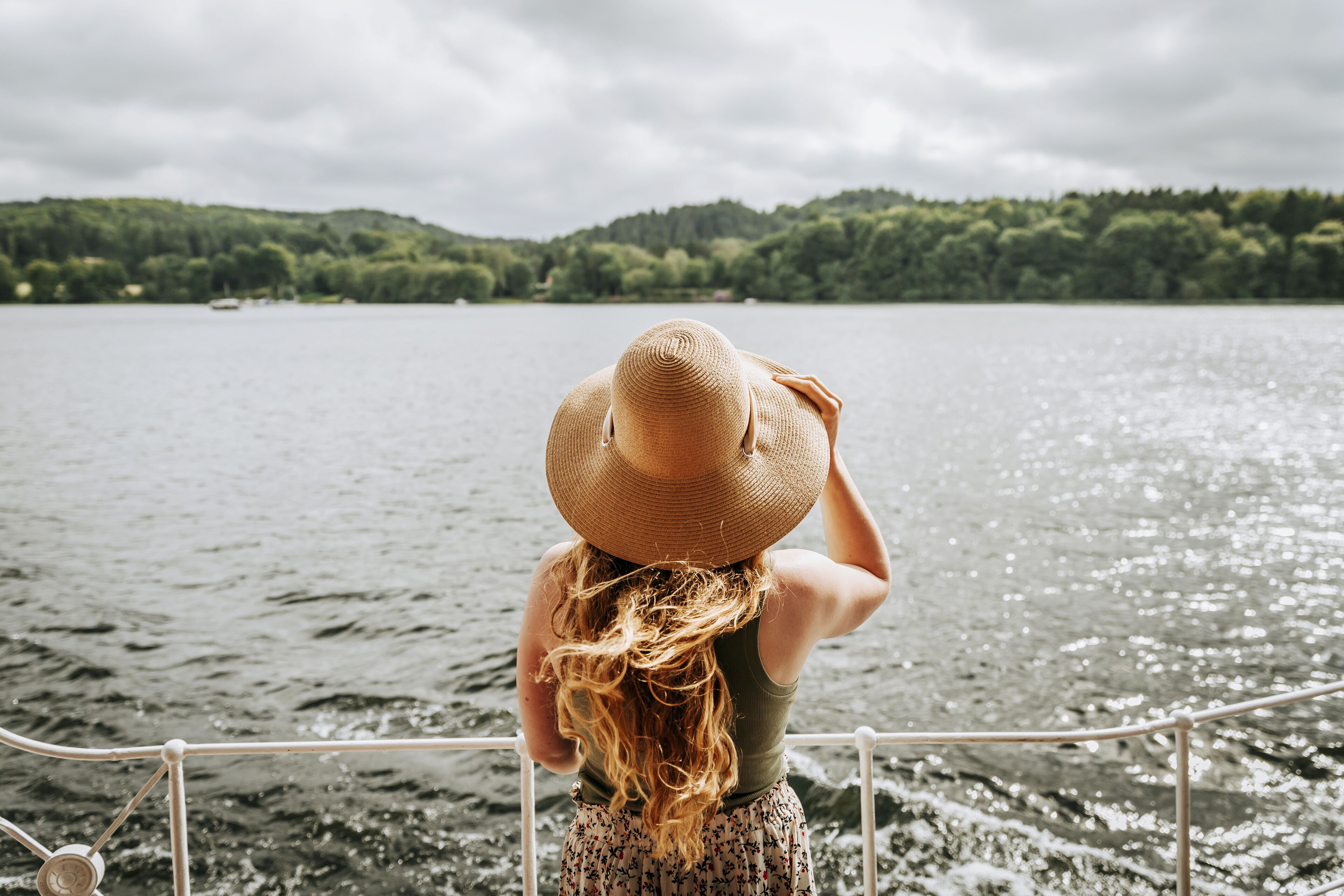 A person with long hair wearing a straw hat, standing by a railing overlooking a body of water with a forested hill in the background.