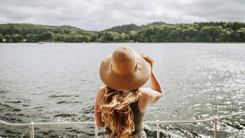 A person with long hair wearing a straw hat, standing by a railing overlooking a body of water with a forested hill in the background.