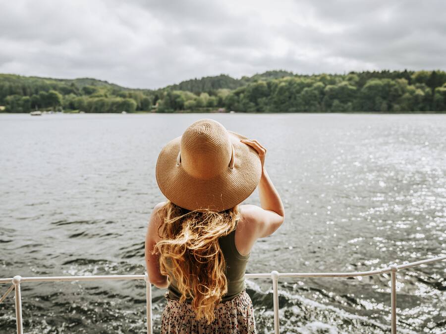 A person with long hair wearing a straw hat, standing by a railing overlooking a body of water with a forested hill in the background.