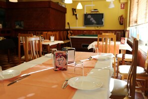 A restaurant with tables set for dining, wooden chairs, a pool table, and a television on the wall.
