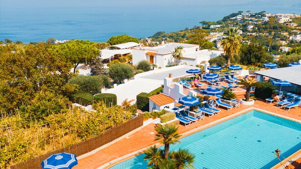 Piscina de un resort con sombrillas azules y sillas de descanso, rodeada de follaje y edificios.