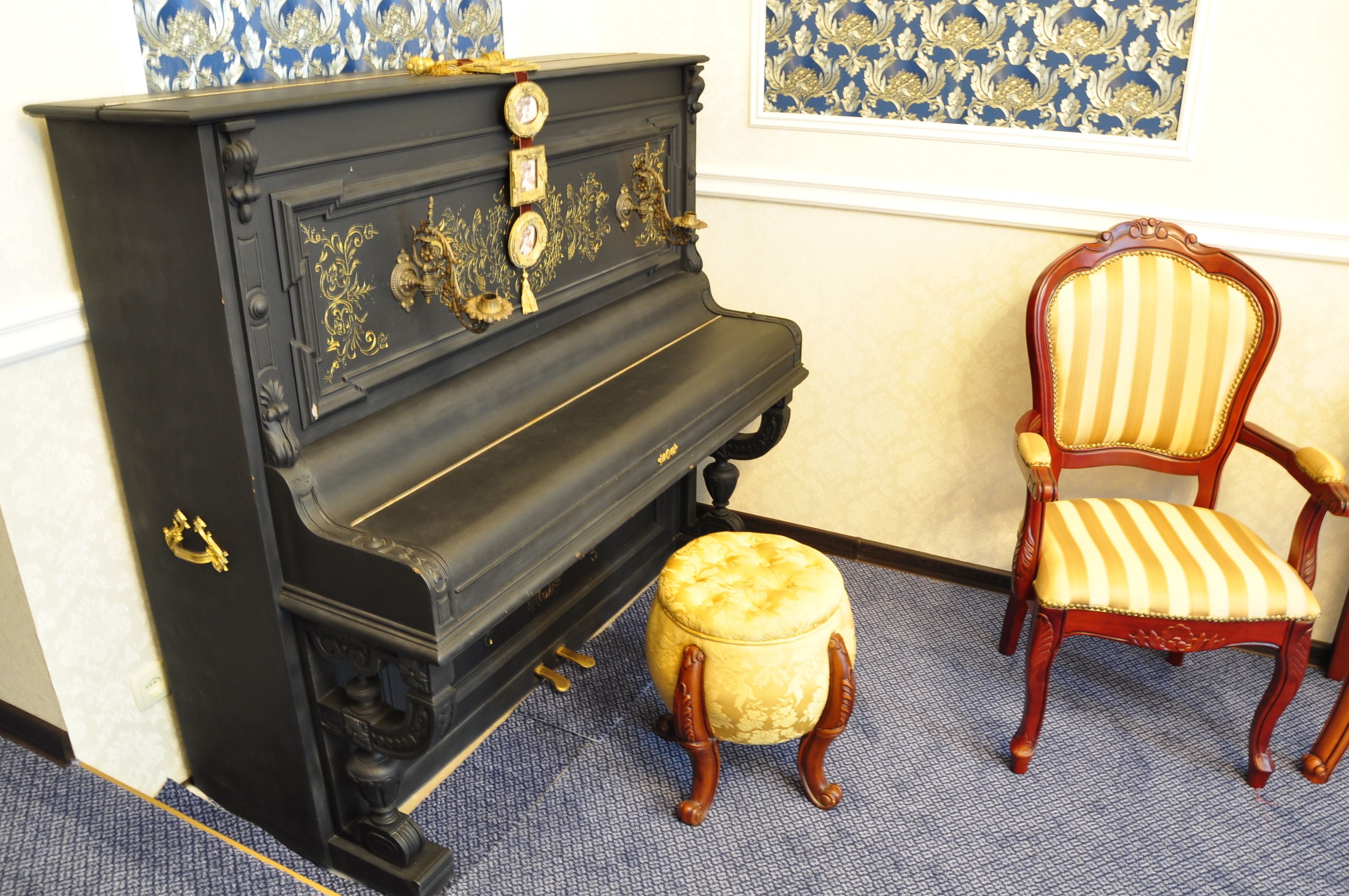 A black piano with gold accents, a matching ottoman, and a striped chair in a room with patterned wallpaper.