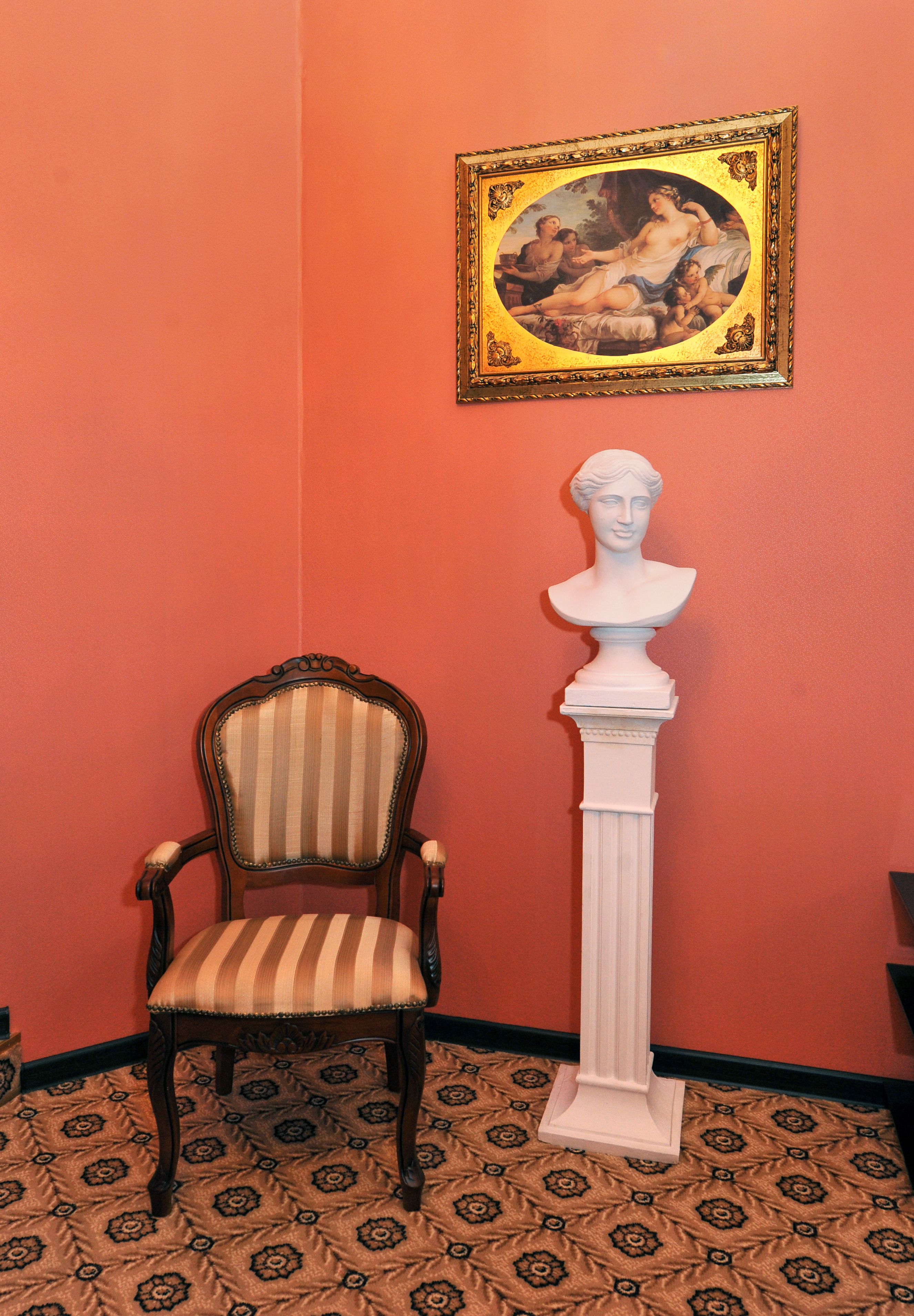 Una habitación con alfombra estampada, una silla a rayas, un busto sobre un pedestal y un cuadro enmarcado en la pared.
