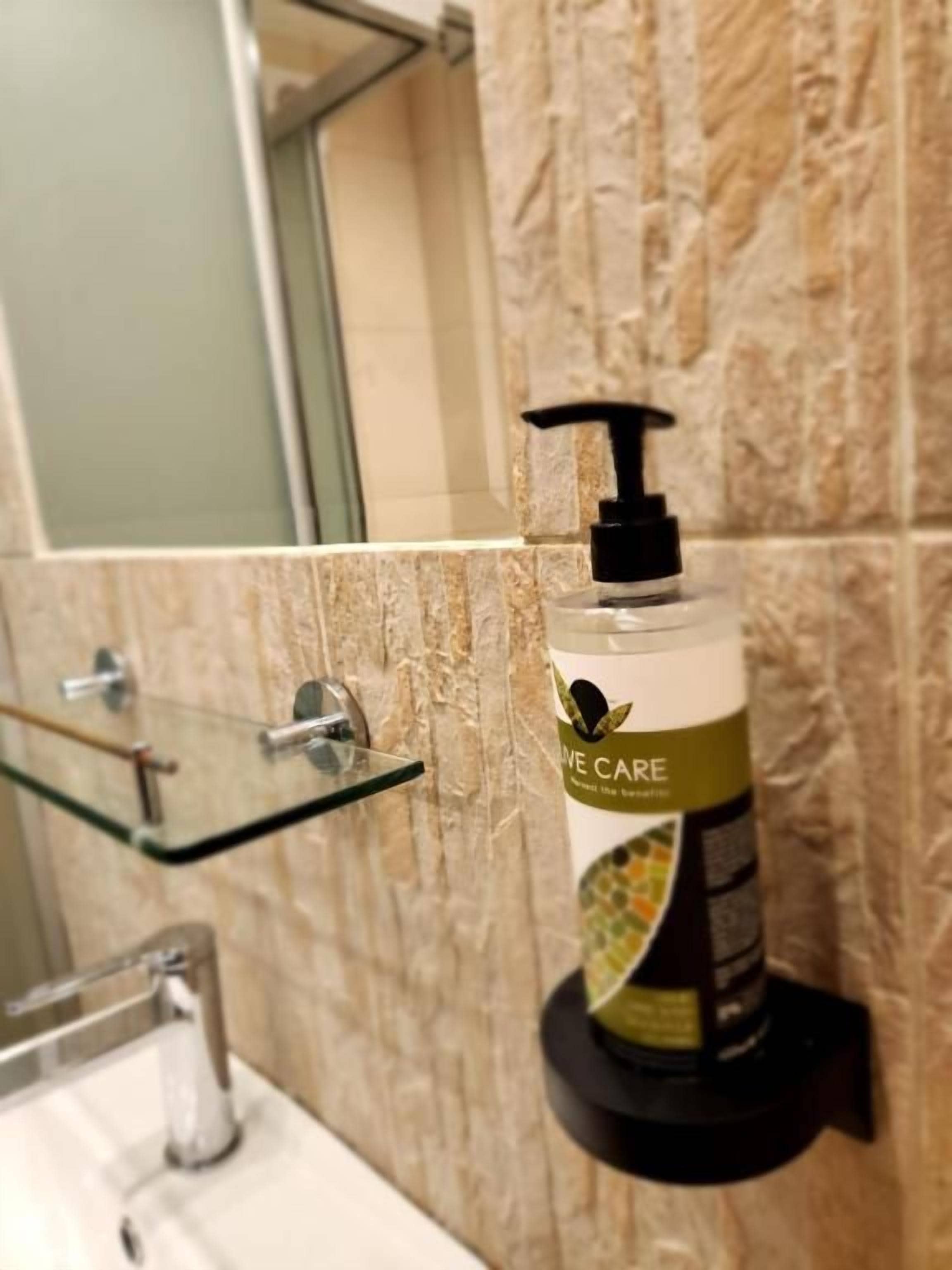 Une salle de bain avec un mur en marbre, une étagère en verre, un distributeur de savon noir et un lavabo blanc.