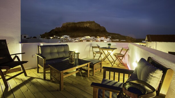 A wooden deck with outdoor furniture, including a sofa, chairs, and a table, overlooking a town with buildings and a hill in the background. - Lindos Boutique Rooms - Adults Only (Rhodes)