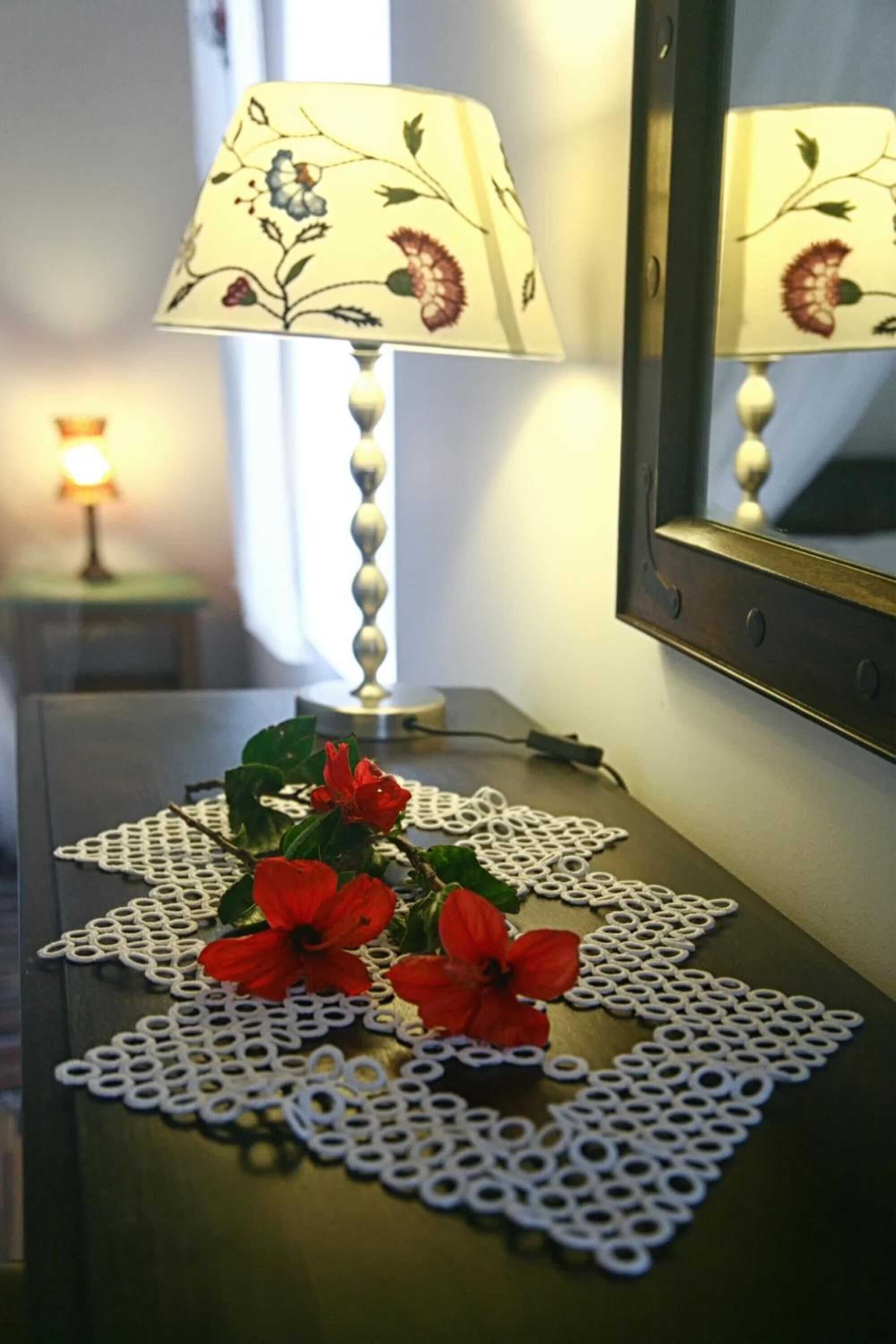 Ein Nachttisch mit Lampe, Spiegel und einer Tischdecke mit roten Blumen.