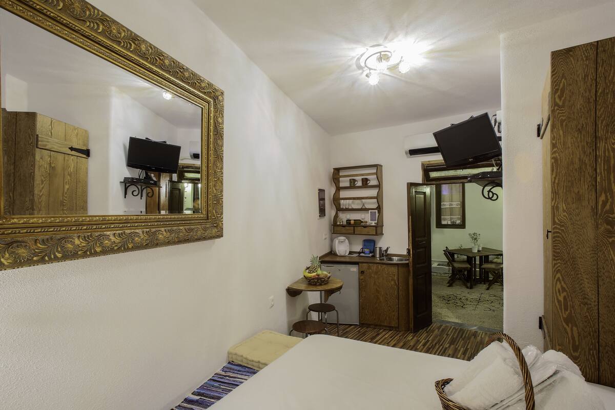 A compact hotel room with a large mirror, a bed, a small kitchenette, and a dining area visible through an open door.