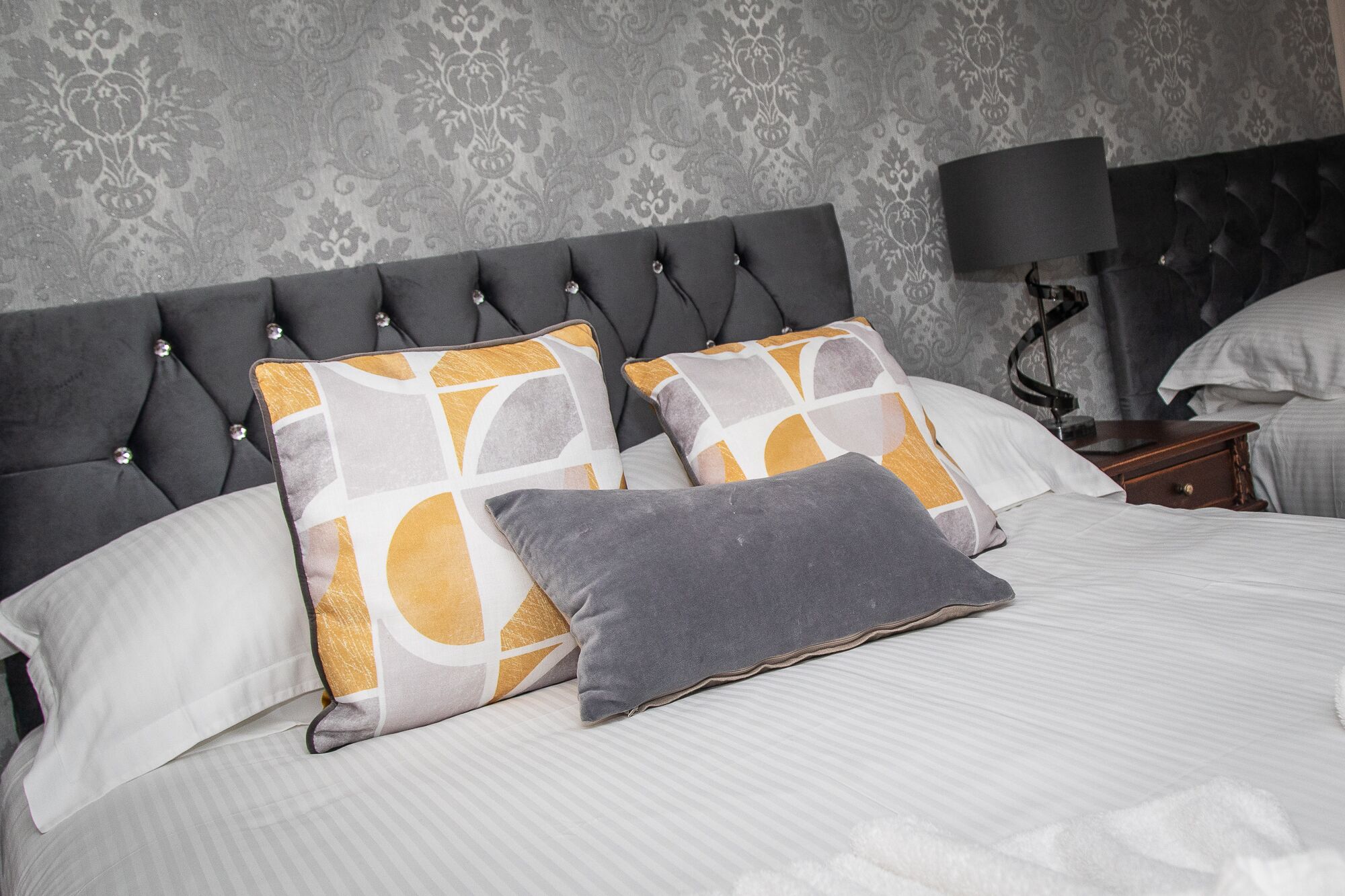 Una cama bien hecha con un cabecero gris, dos almohadas estampadas y un cojín gris.