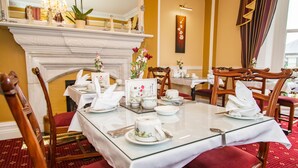 Un comedor con una mesa de vidrio, sillas tapizadas rojas, manteles blancos y una chimenea con adornos.