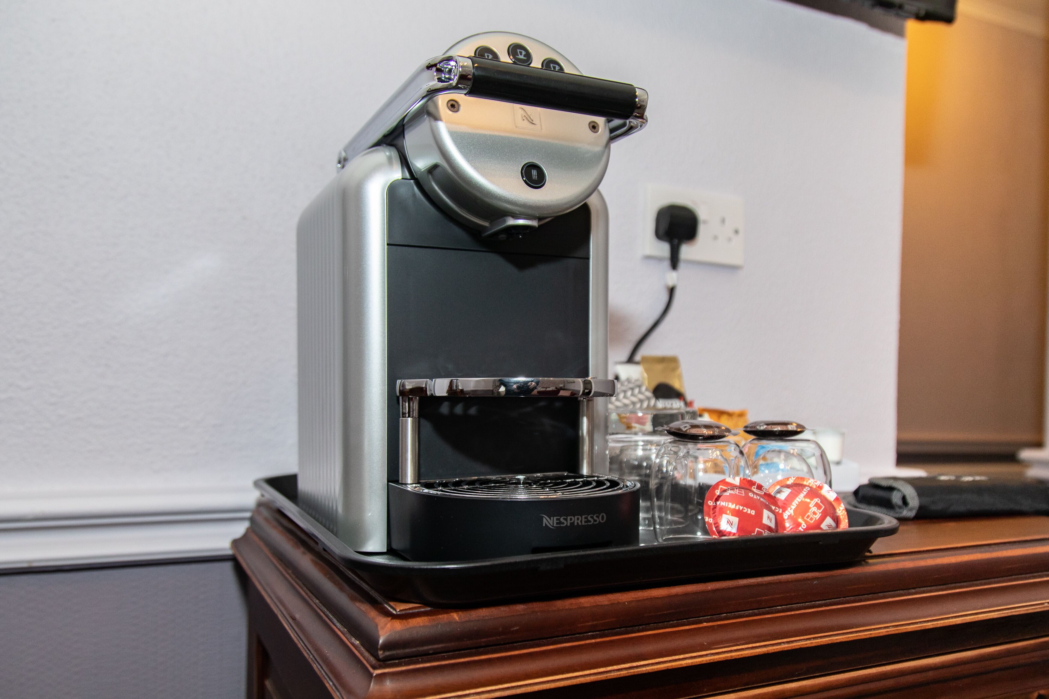 Une machine à café Nespresso posée sur un comptoir en bois, avec un plateau de capsules de café et une carafe en verre.