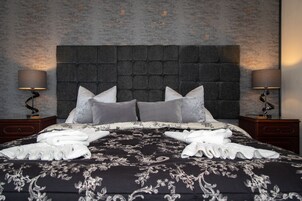 Una cama bien tendida con una colcha oscura estampada y almohadas blancas, flanqueada por dos mesitas de noche con lámparas, frente a una pared con textura.