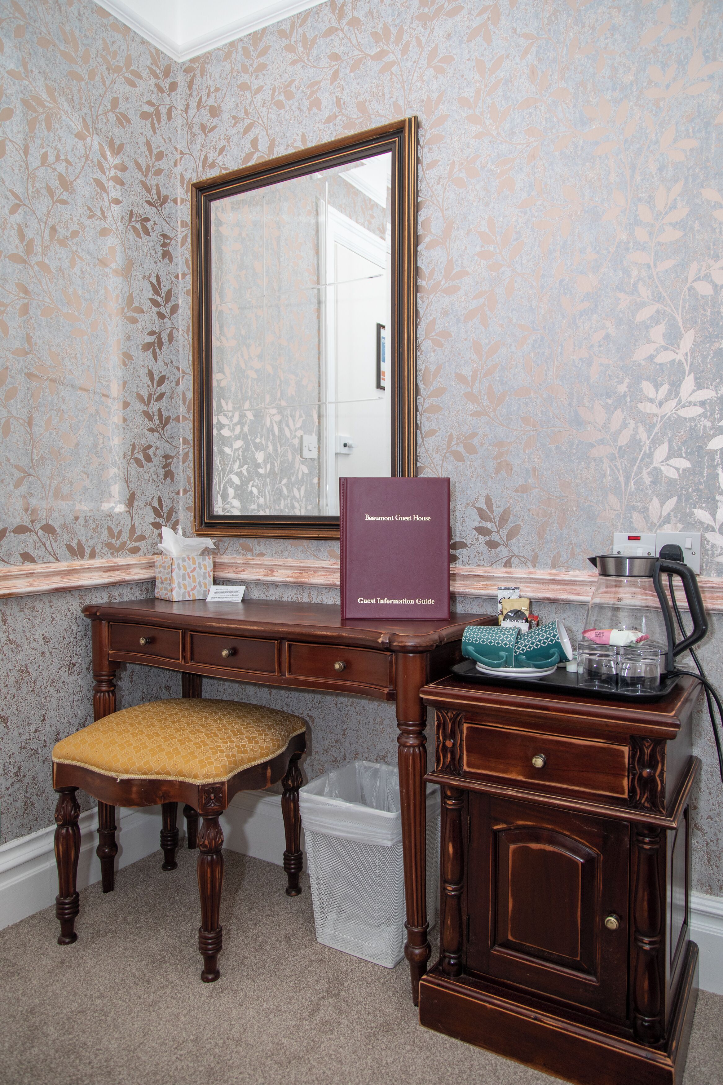 A room with a wooden desk, a mirror, a guest information guide, a small table with a coffee maker, and a trash bin.