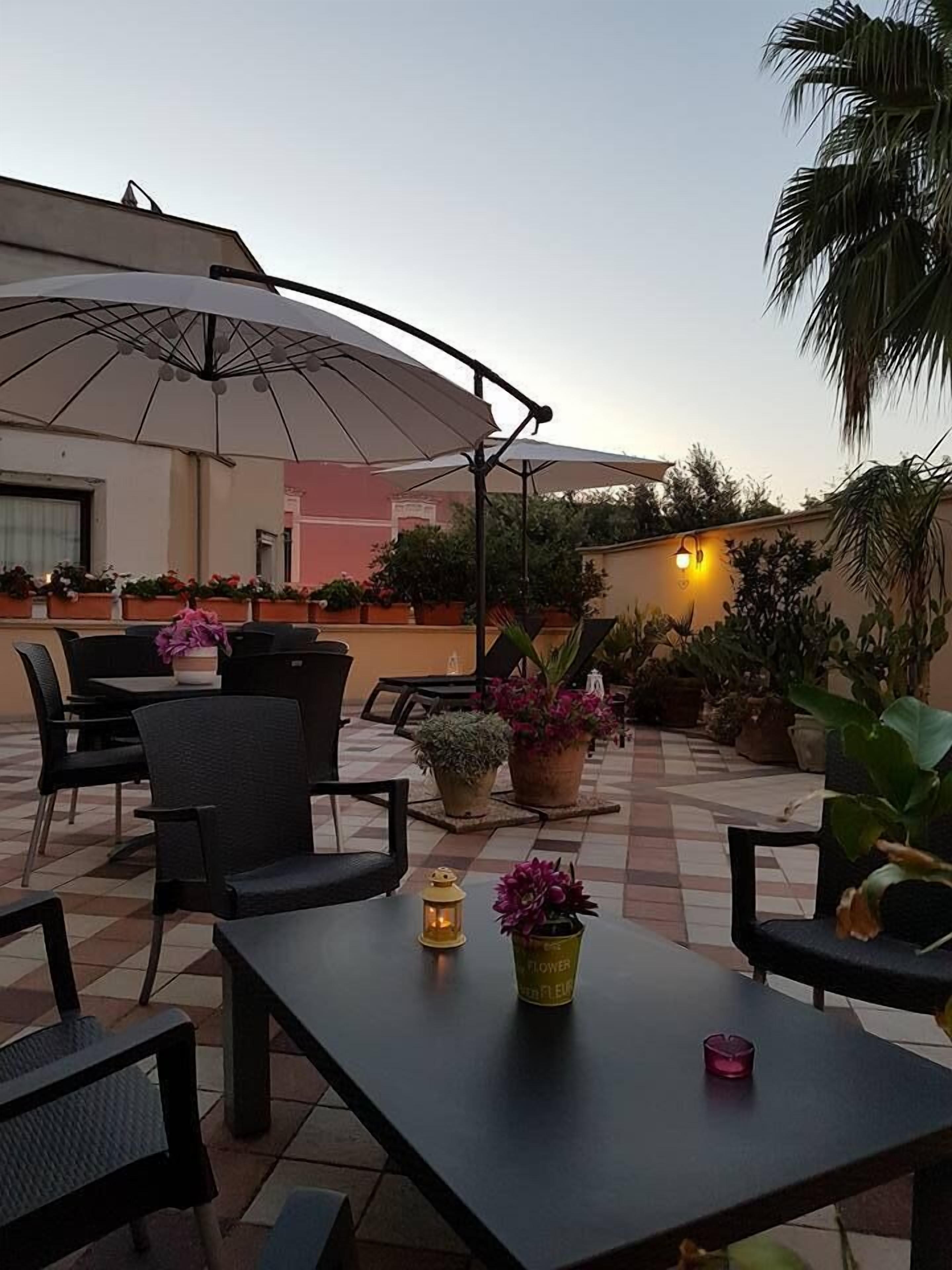 An outdoor patio area with tables and chairs, umbrellas, potted plants, and a tiled floor.