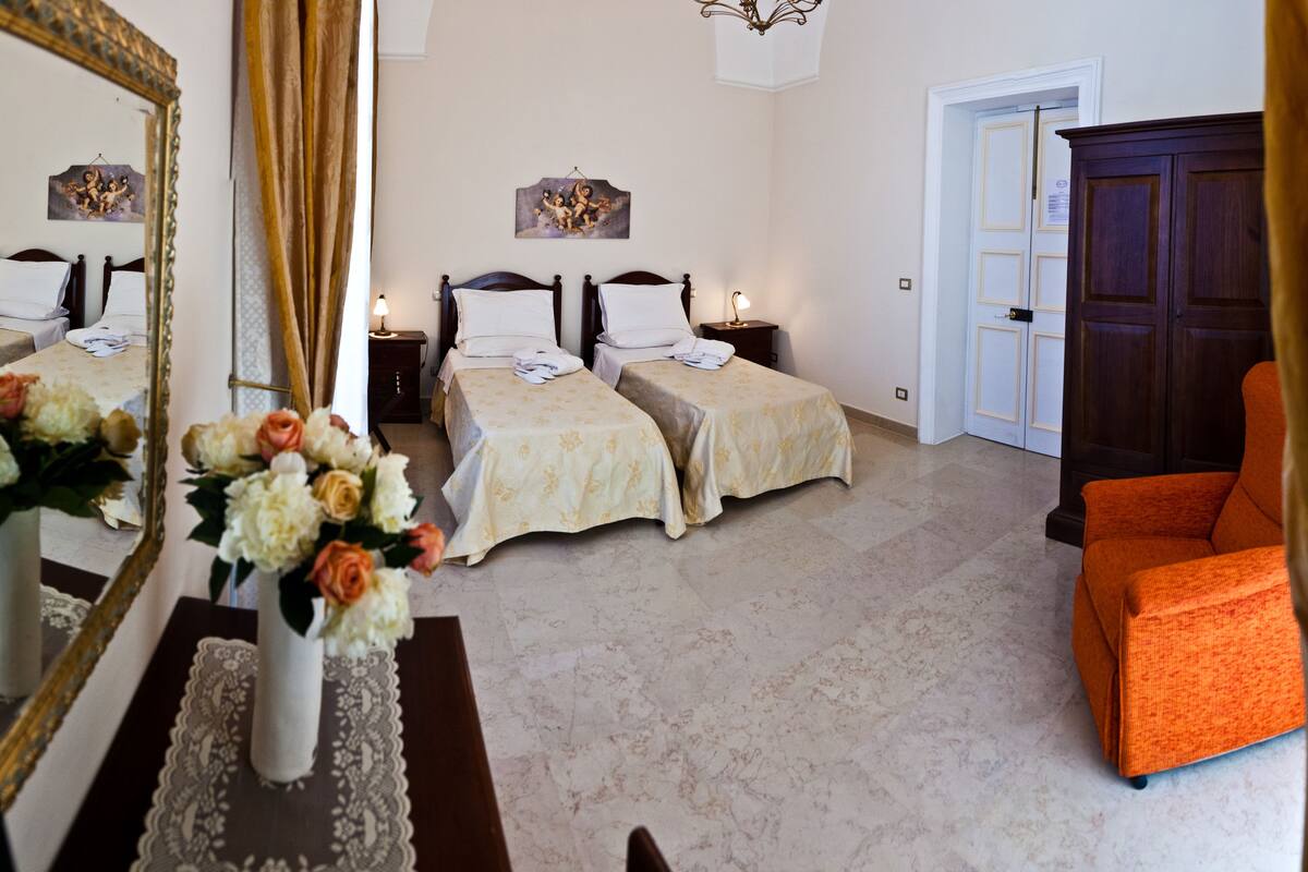 Une chambre d’hôtel avec deux lits, un grand miroir, un vase avec des fleurs et une porte donnant sur une autre pièce.