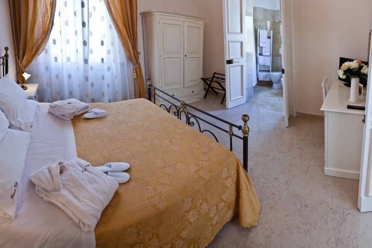 Un lit bien fait, avec du linge de lit blanc et une couvre-lit dorée, dans une pièce où se trouvent une armoire blanche, une petite table et une chaise.