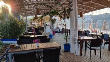 An outdoor dining area with wooden tables and chairs, surrounded by potted plants and umbrellas.