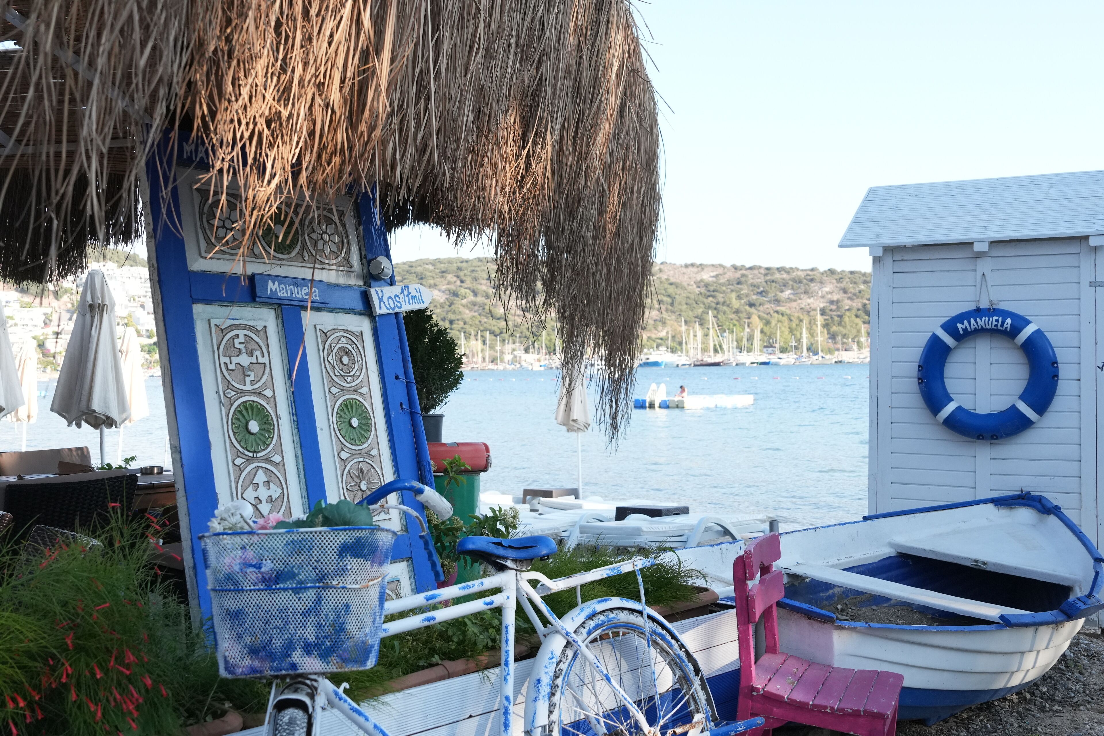 A beachside scene with a blue and white building labeled 'Manuela' and a small boat docked nearby.