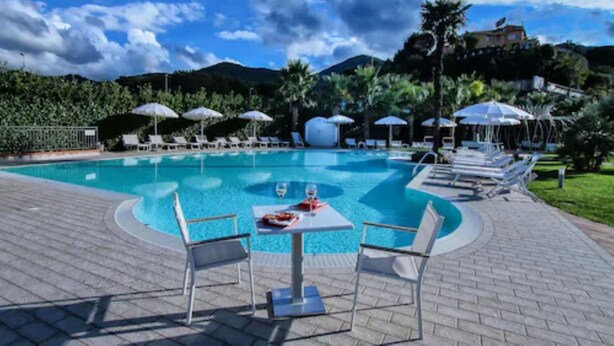 Zona de piscina con sillones de descanso y sombrillas, rodeada de vegetación y colinas.